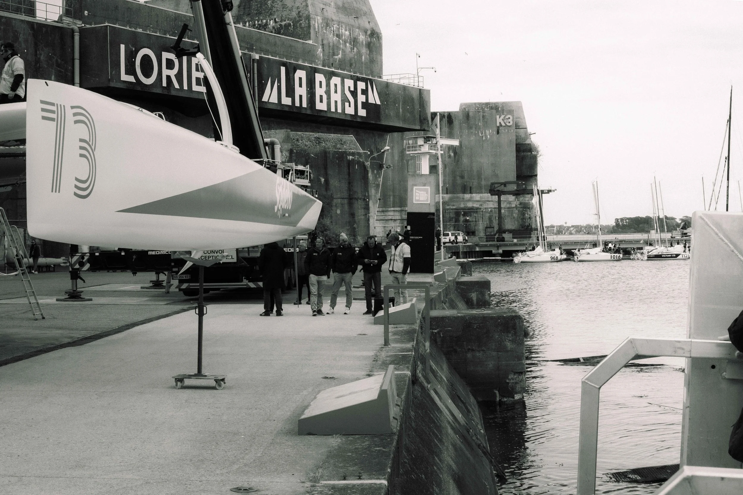 Vue d'un port avec un quai, plusieurs voiliers amarrés, un bateau sur le quai avec des hommes en discussion, un grand mur en béton avec des inscriptions, et des lampadaires.