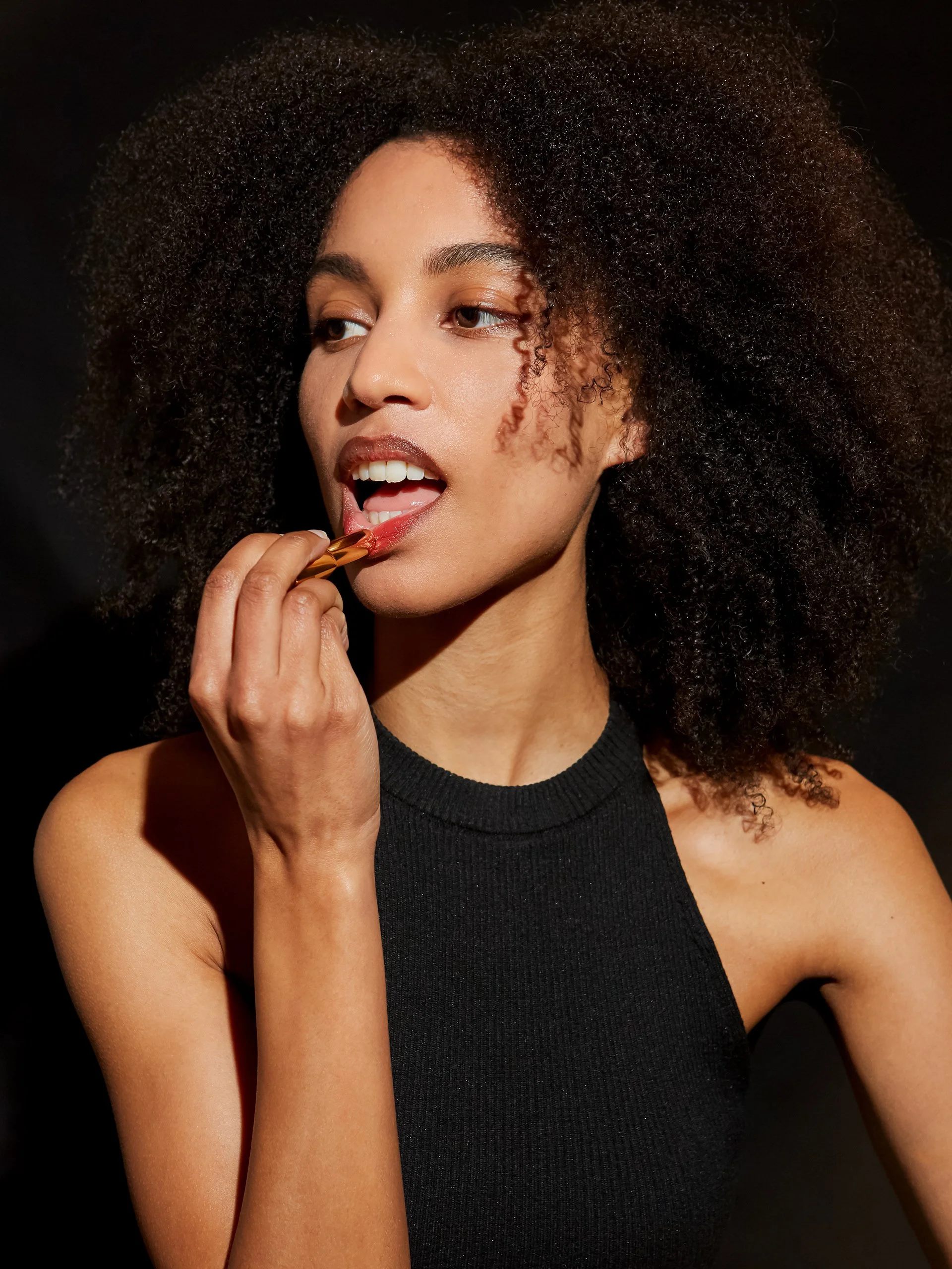 une femme avec de longs cheveux bouclés et une robe noire, qui se maquille ou se prépare, contre un fond noir