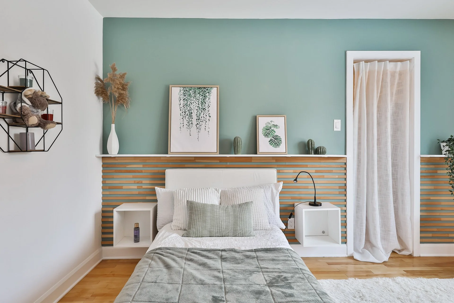 Chambre moderne avec un lit face à un mur vert avec un motif en bois. Décor avec deux tableaux, une grande vase avec des plantes séchées, et trois petits poteries. Étagère murale en forme de géométrie avec des objets. Une lampe de lecture simple sur 
