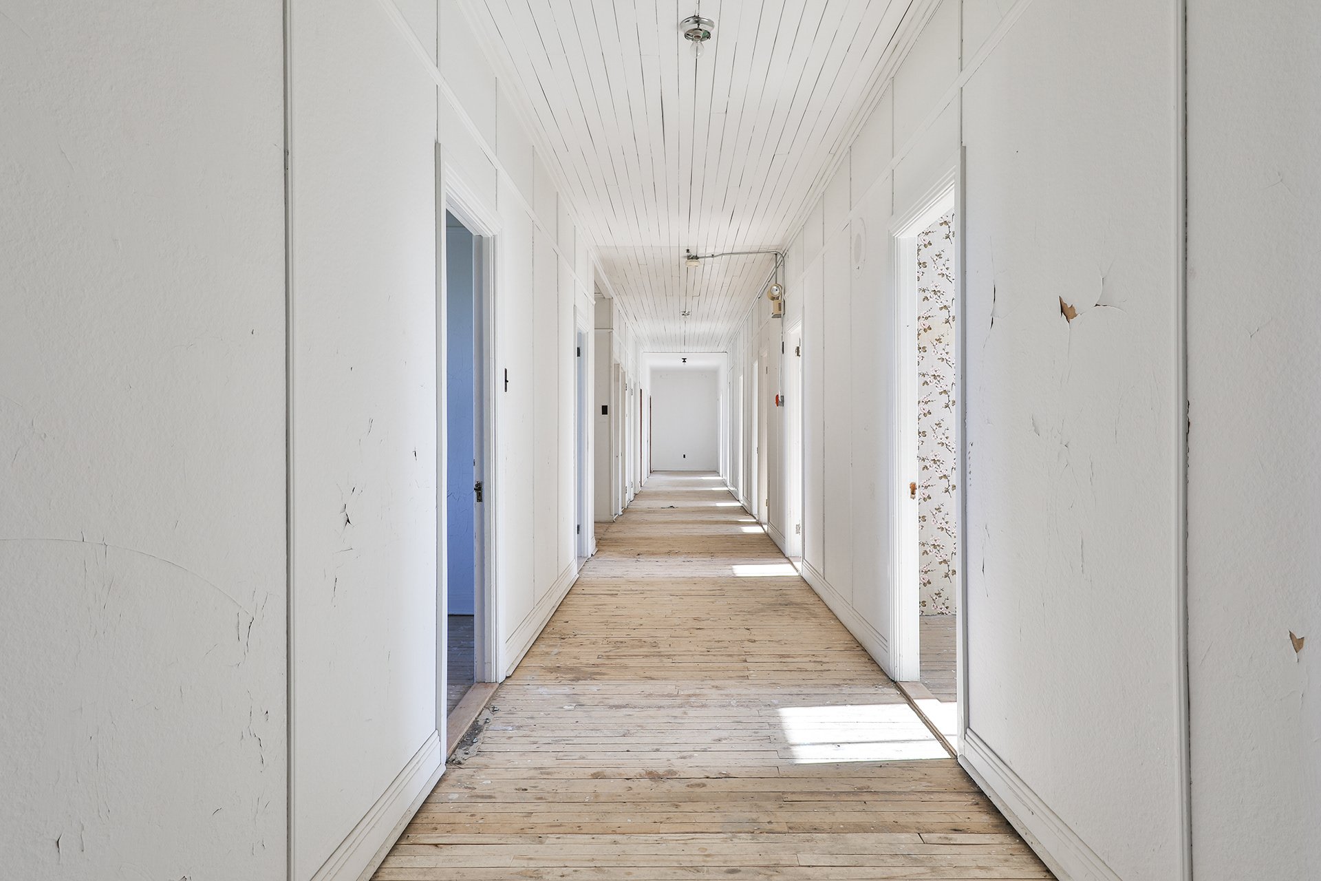 Couloir intérieur avec murs blancs, sol en bois brut, et plusieurs portes ouvertes. La lumière naturelle éclaire la scène.