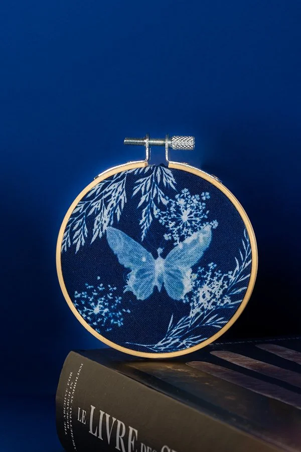 Infernière image d'une pièce de broderie avec un papillon bleu au centre, entouré de motifs de feuilles et de fleurs, fixé dans un cadre à broder.