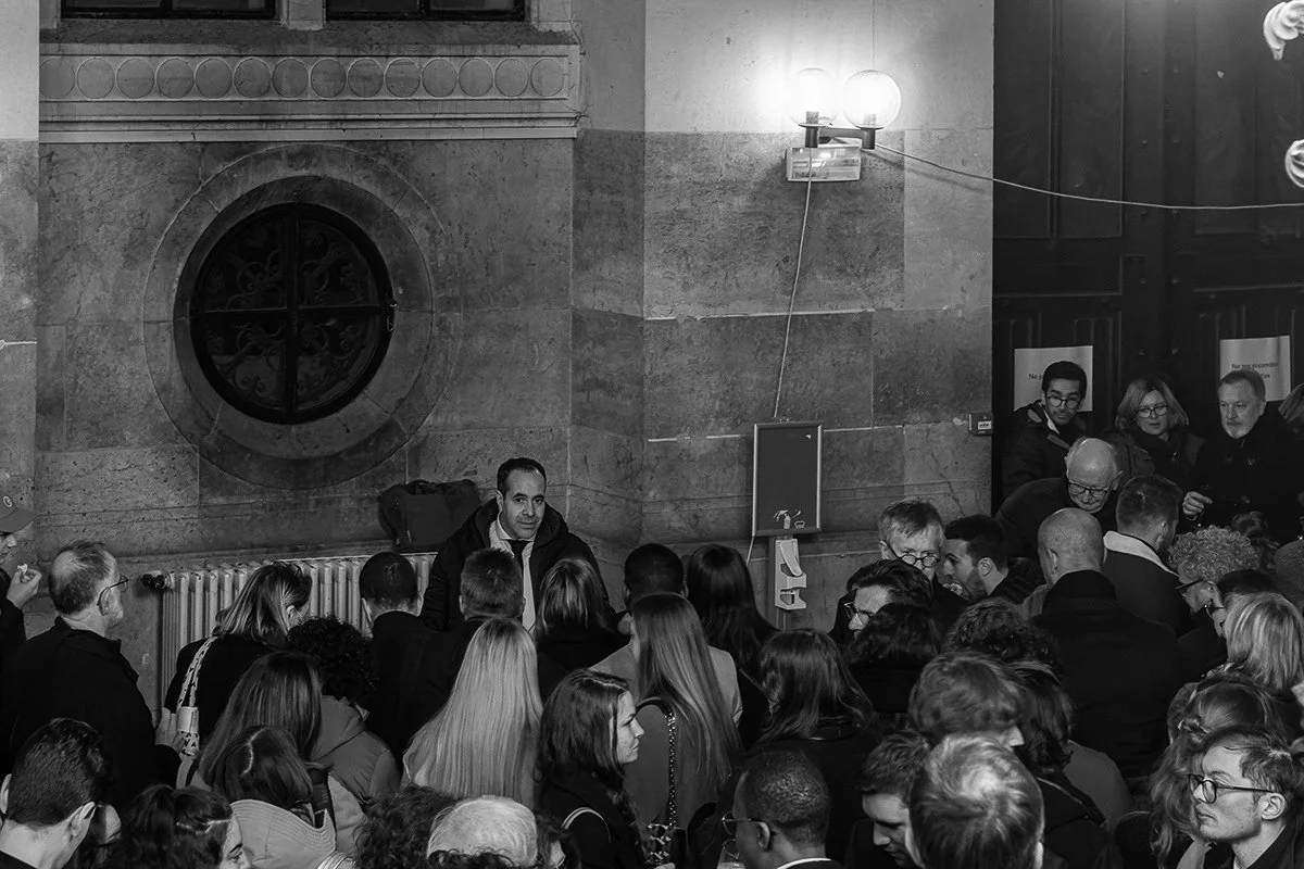 Un groupe de personnes rassemblées dans une pièce, avec un homme au centre semblant parler ou répondre.