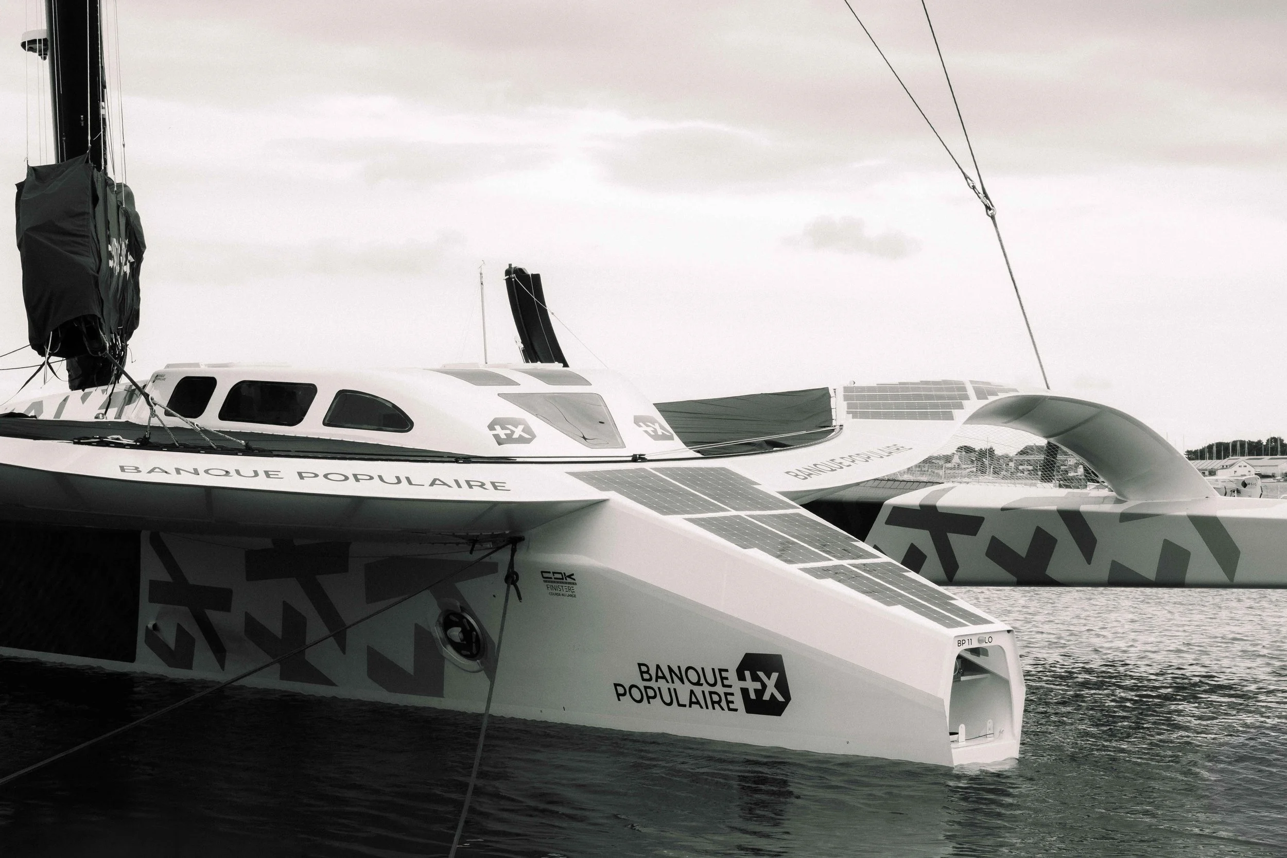 Deux bateaux à voile solaires avec l'inscription 'BANQUE POPULAIRE' et le logo de la banque, flottant sur l'eau dans un port, avec un ciel partiellement nuageux.
