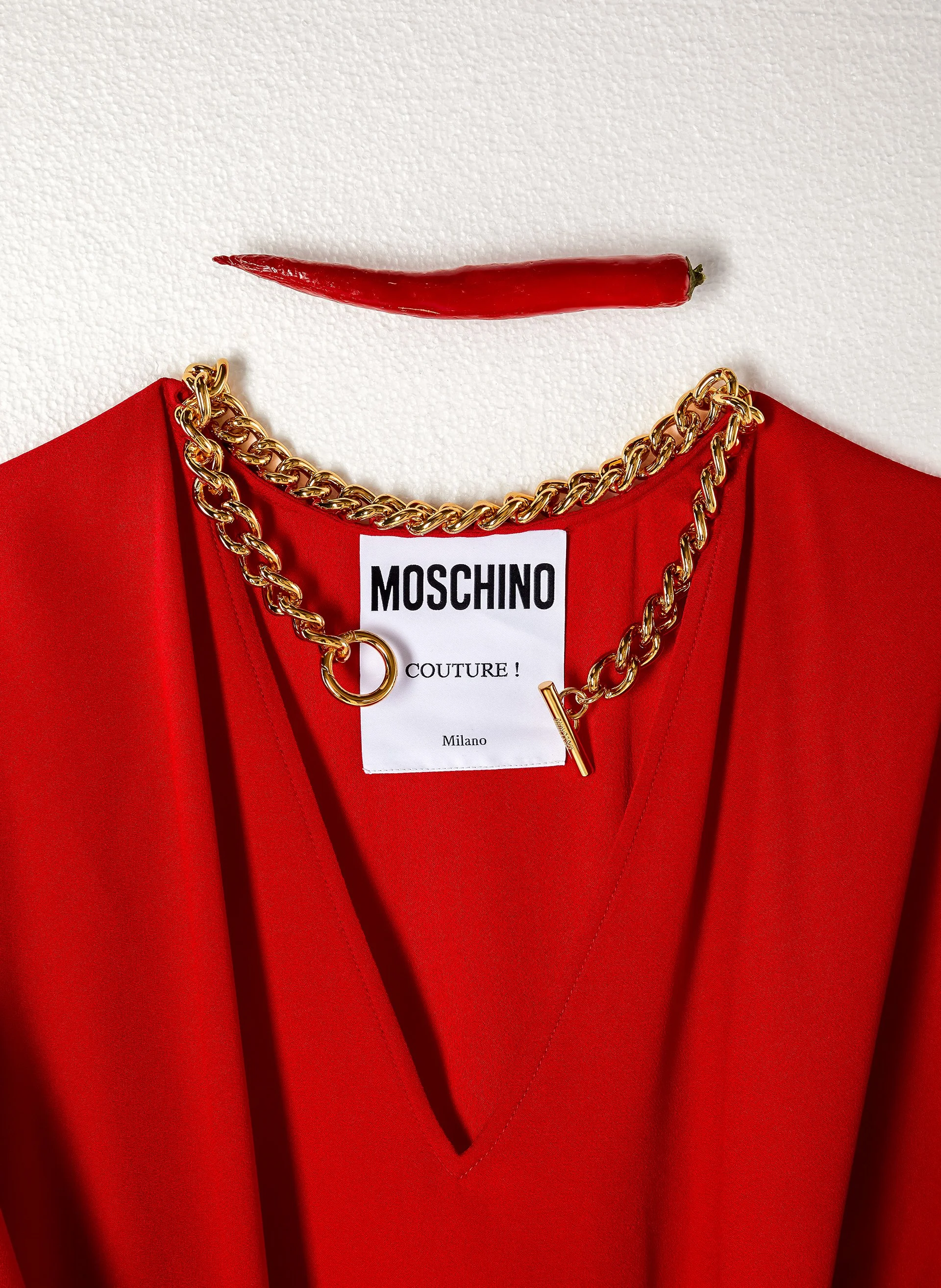 Une robe rouge avec une étiquette de la marque Moschino, un collier en chaîne dorée et un piment rouge au-dessus.