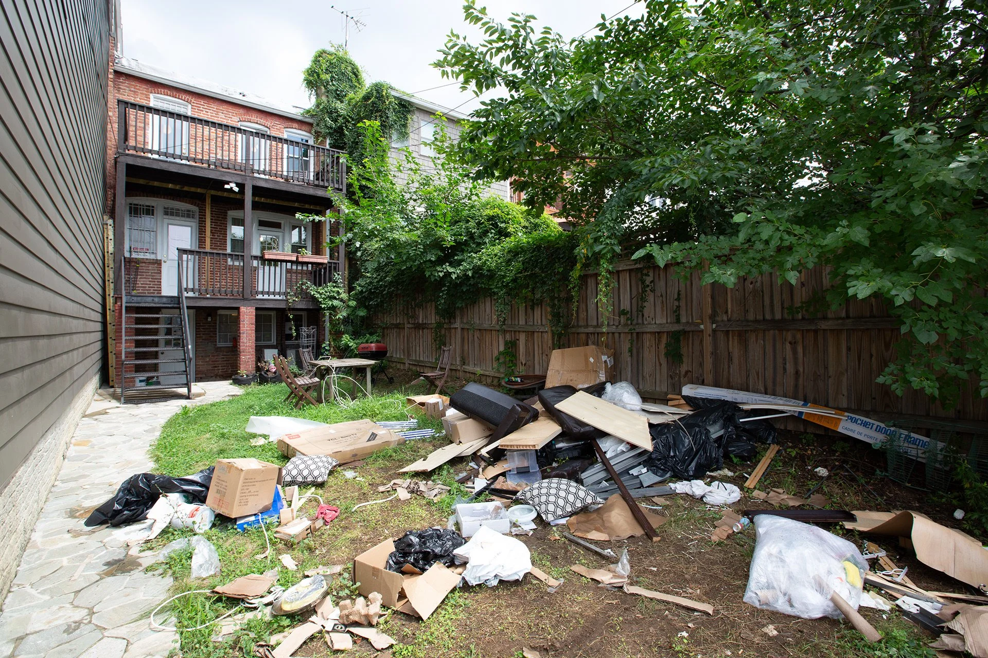 Jardin résidentiel avec déchets, cartons, plastique et bois en désordre, entouré de haies et clôture en bois, avec un bâtiment à plusieurs étages en arrière-plan.