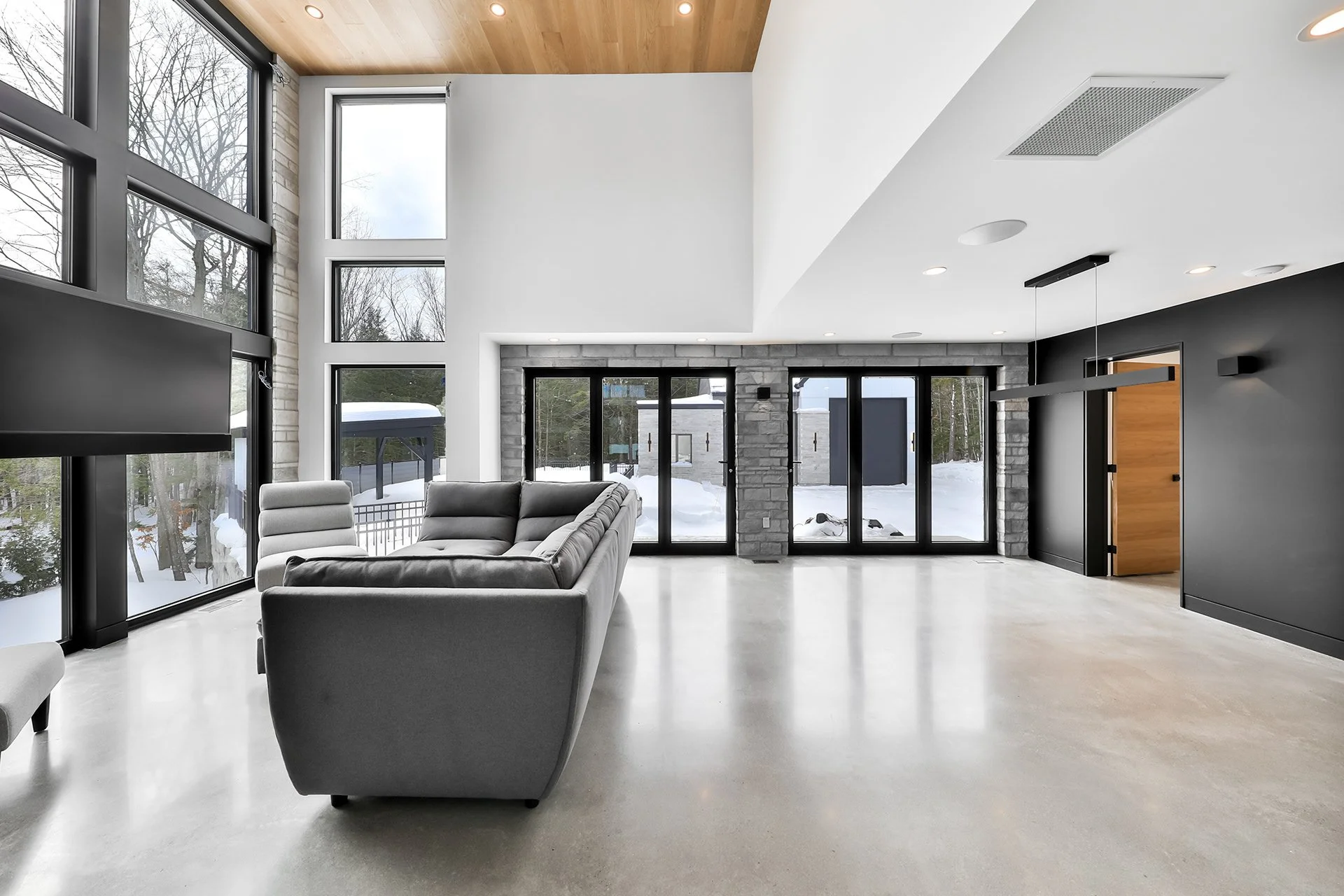 Salon moderne avec grands fenêtres, canapé gris, sol en béton, murs peints en blanc et noir, porte en bois, vue sur extérieur enneigé.