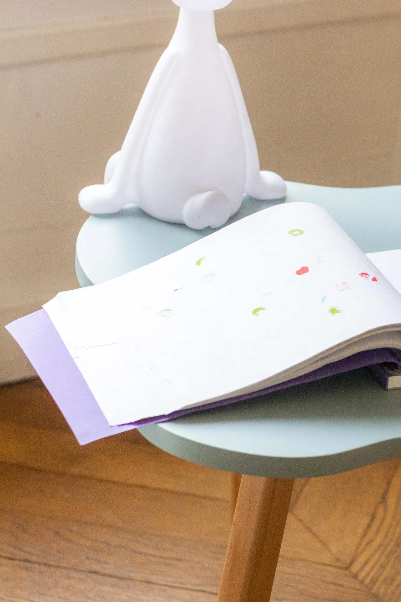 Un livre ouvert posé sur une petite table arrondie, avec une figurine blanche d'animal à quatre pattes à côté, dans un environnement intérieur.