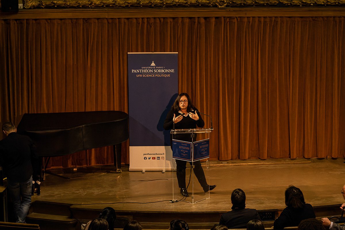 Une femme parle à un micro sur scène avec une bannière de l'Université Paris 1 Panthéon Sorbonne en arrière-plan, devant un auditoire. Il y a un piano sur le côté gauche.