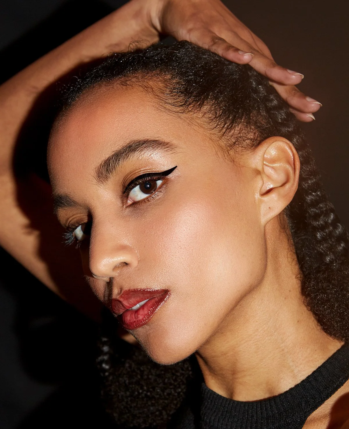 Jeune femme avec maquillage, portant un choker noir, pose face à une caméra, main sur la tête, fond sombre.