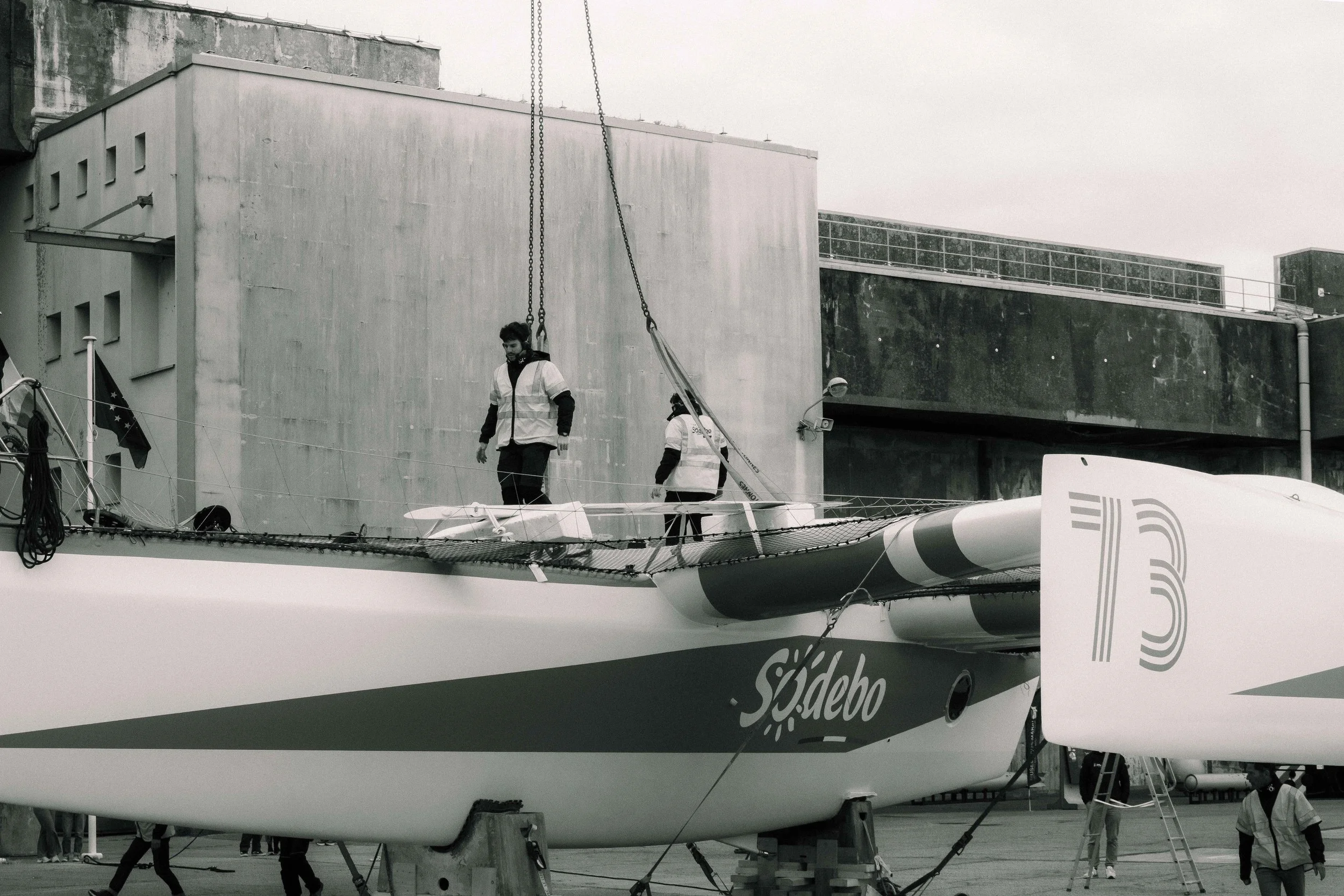Image en noir et blanc d'un bateau de course à voile en préparation, avec des personnes en uniforme sur le pont et à terre.