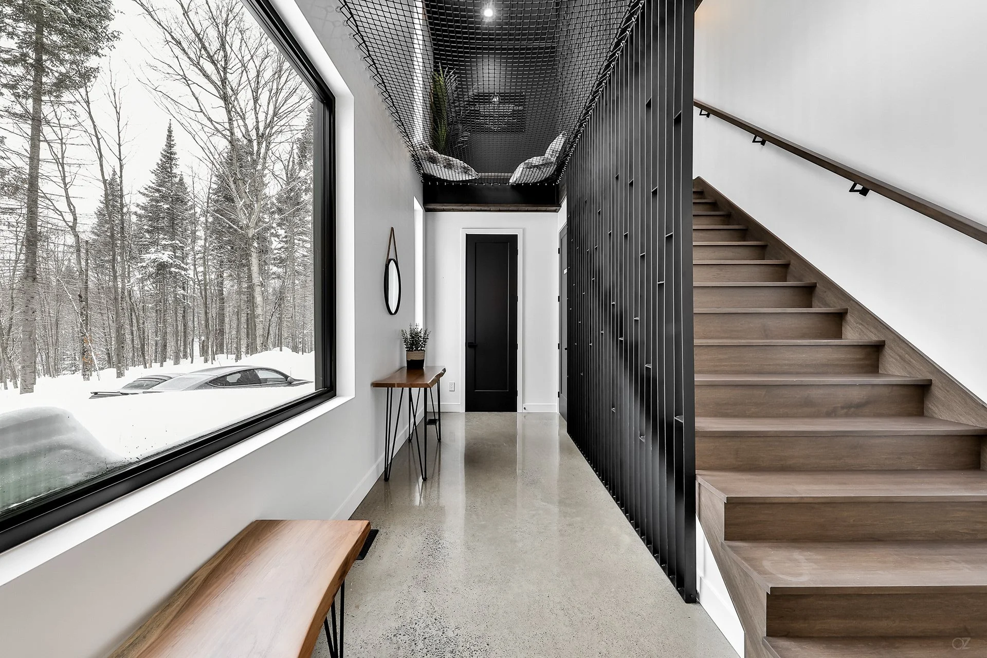 Intérieur moderne d'une maison avec escalier en bois, mur noir, grande fenêtre avec vue sur un paysage enneigé.