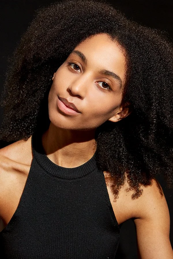 Jeune femme aux cheveux bouclés et noire portant un haut noir, regardant l'appareil avec un fond noir.