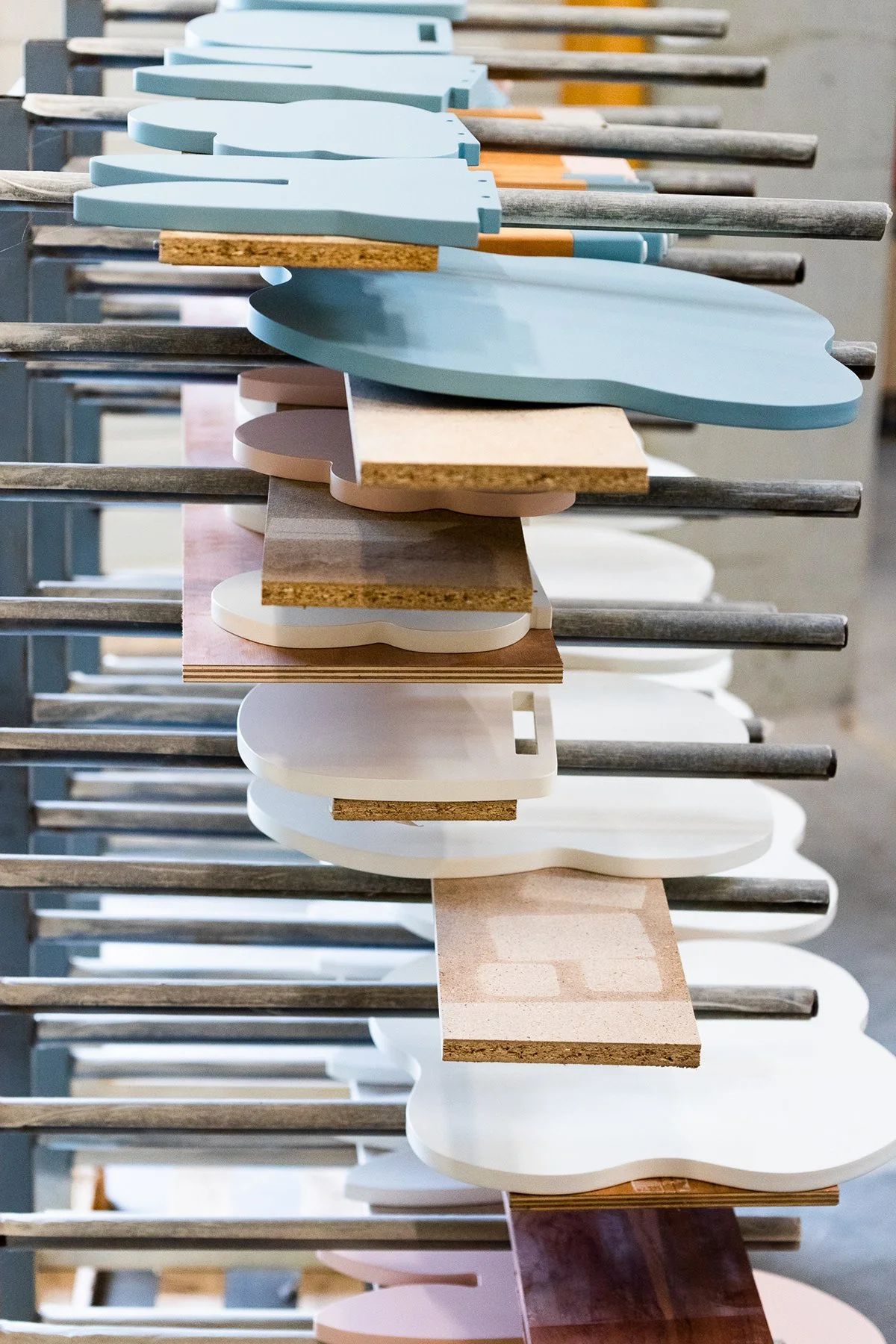 Une série de panneaux en bois peint, en blanc, beige, bleu clair, et d'autres couleurs, suspendus horizontalement sur une structure métallique pour la fabrication ou l'assemblage.