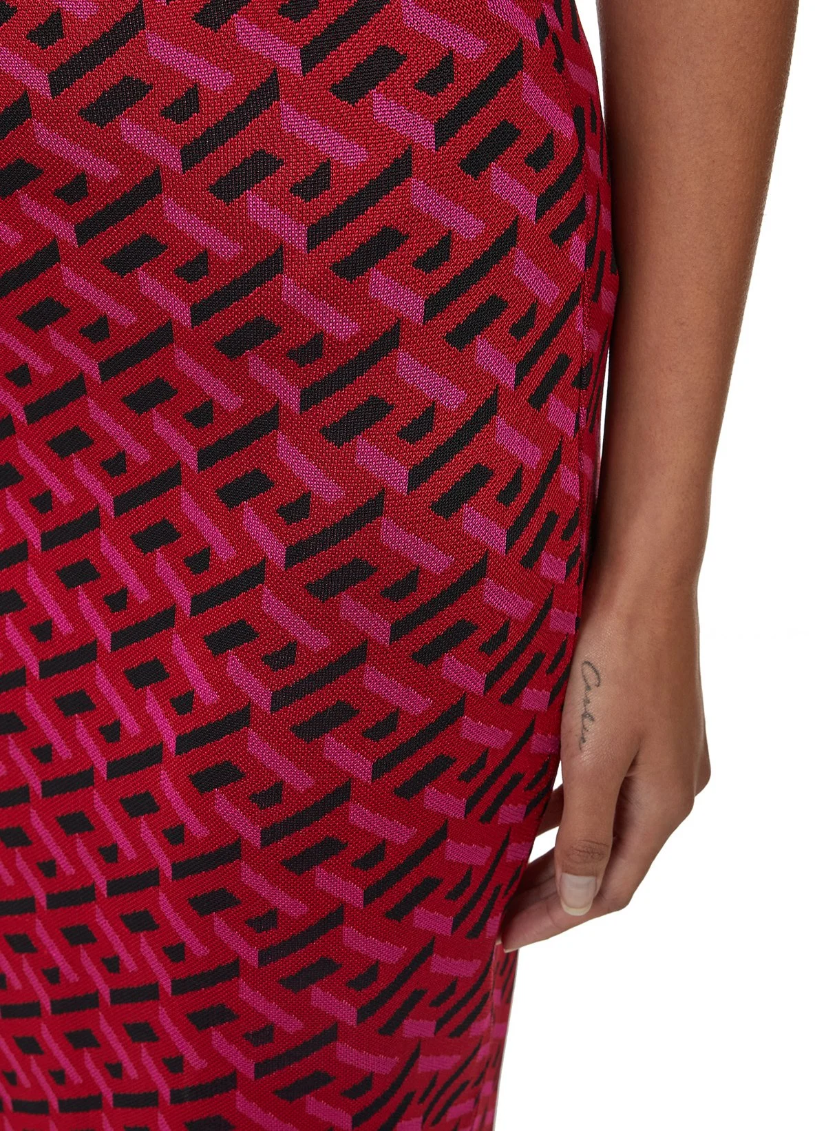 Une personne porte une robe avec un motif géométrique rouge, noir et violet, et a un tatouage 'Calva' sur le doigt.