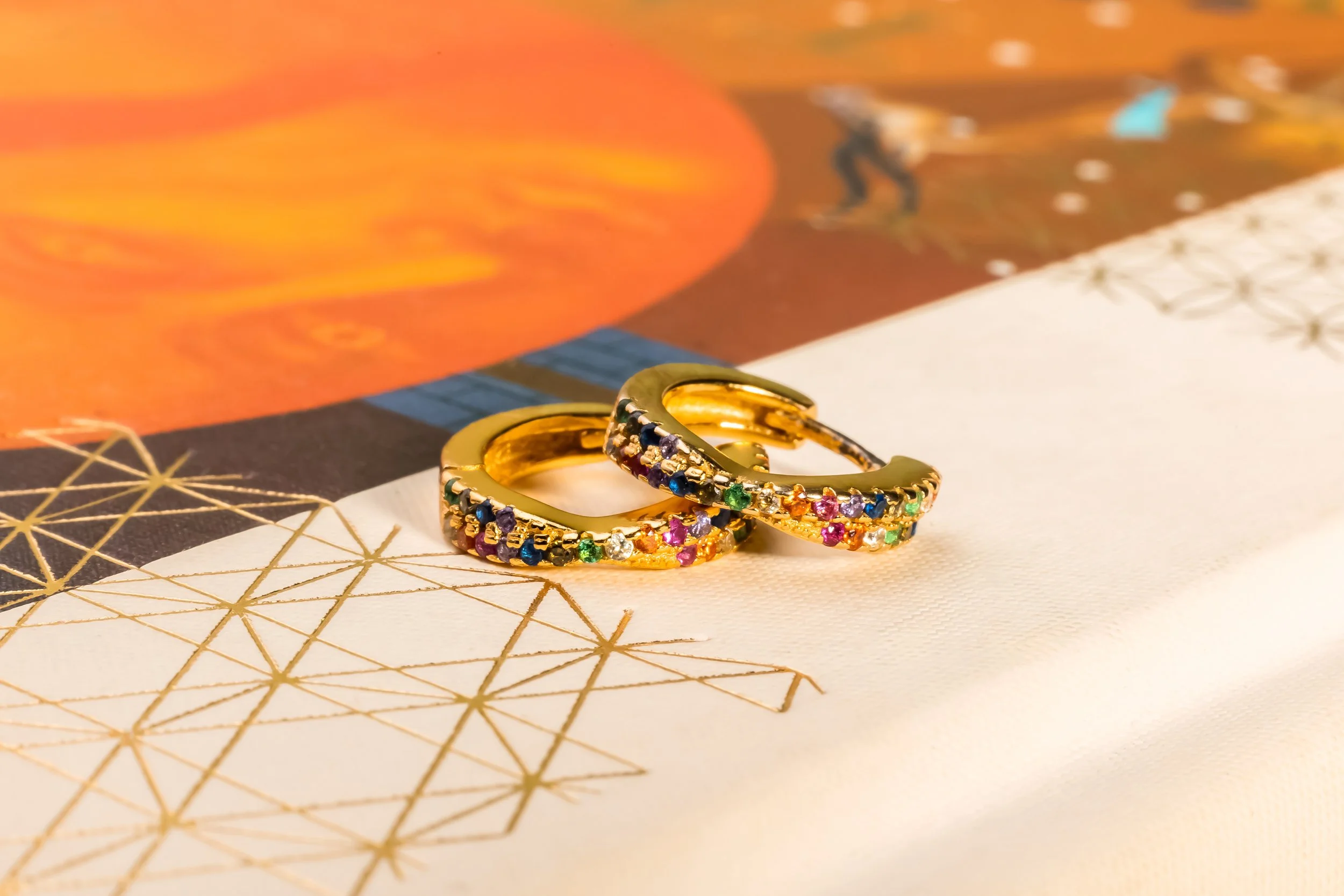 Deux alliances en or ornées de pierres précieuses colorées, posées sur un fond blanc avec un papier décoratif et une image illustrée en arrière-plan.