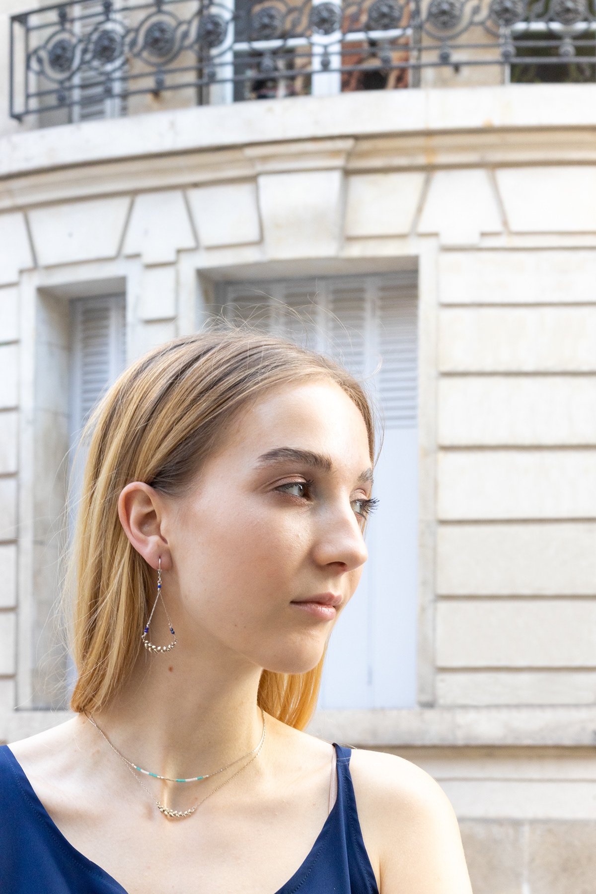 Jeune femme aux cheveux blonds portant un collier et des boucles d'oreilles en outdoor, avec un bâtiment en pierre en arrière-plan.