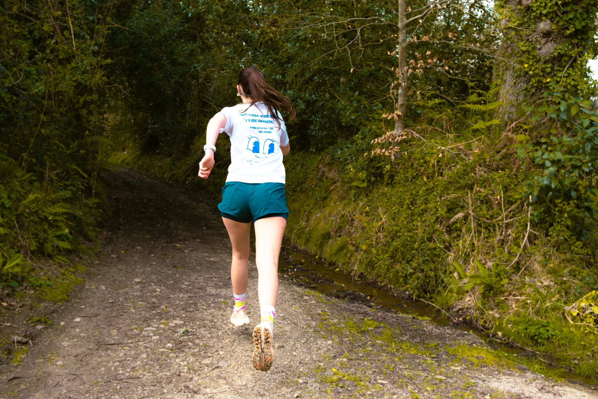 Une femme court sur un sentier en forêt, vue de dos. Elle porte un t-shirt blanc, un short bleu, des chaussettes colorées et des chaussures de course. La végétation verte entoure le chemin.