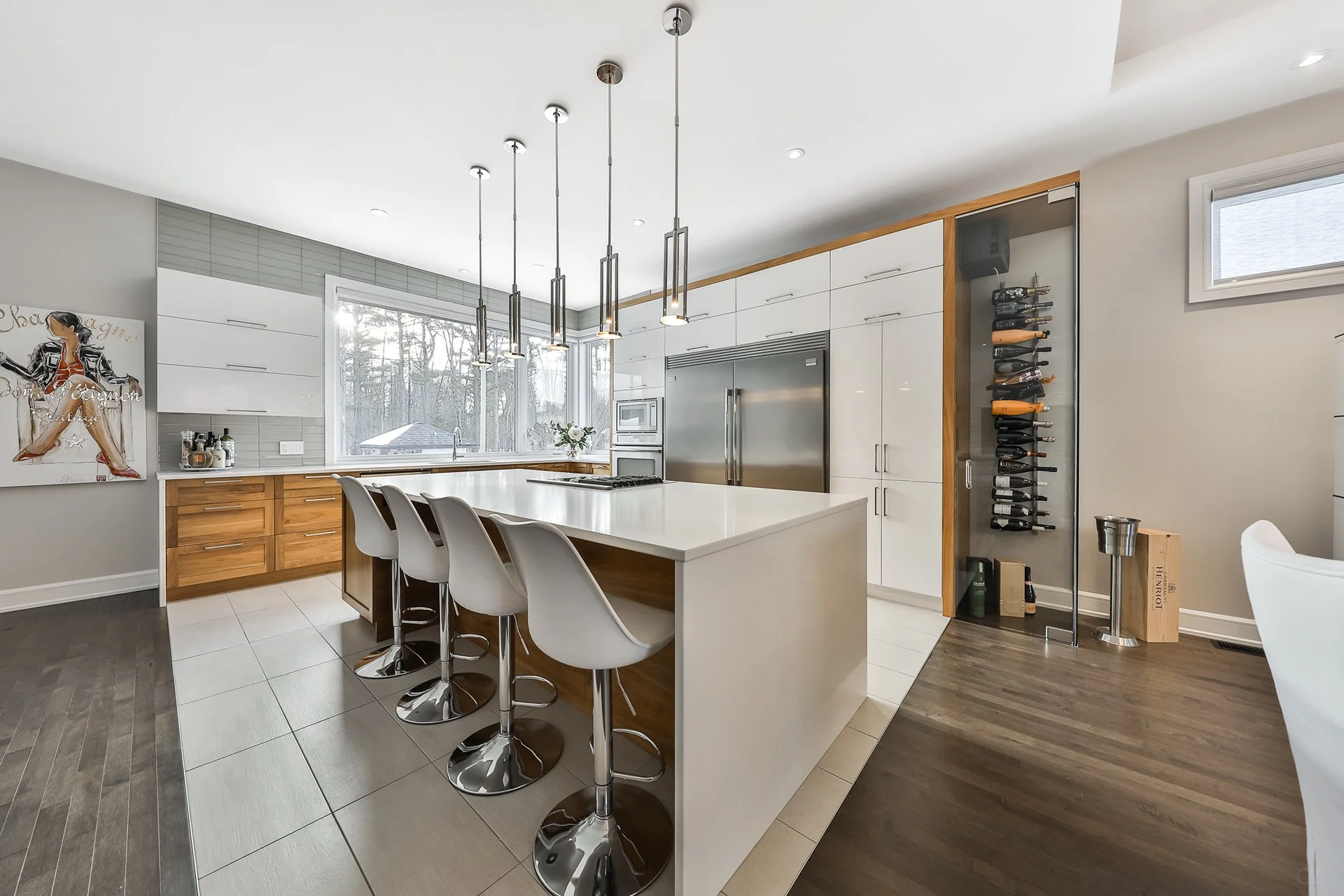 Cuisine moderne avec un grand îlot central, des chaises modernes, des armoires blanches, un réfrigérateur en acier inoxydable, un éclairage suspendu et une grande fenêtre avec vue sur des arbres