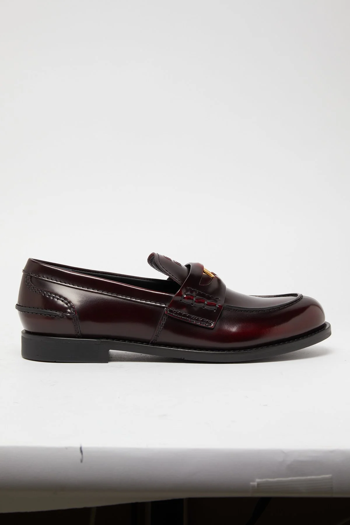 Une chaussure mocassin en cuir marron foncé avec une bride ornée d'une boucle en métal doré, posée sur une surface blanche.