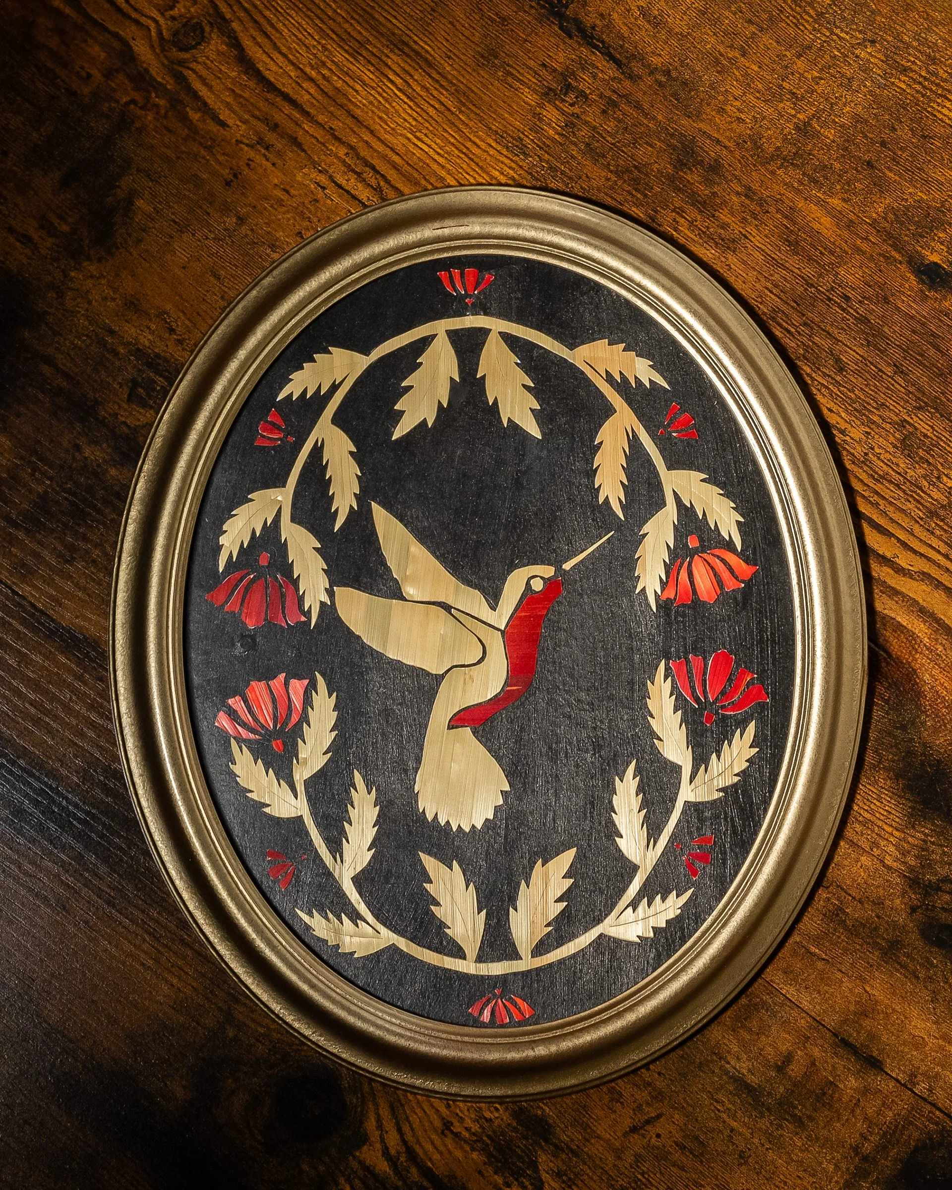 Horloge décorative avec un héron stylisé au centre, entouré de feuilles et de fleurs rouges, dans un cadre ovale en métal.