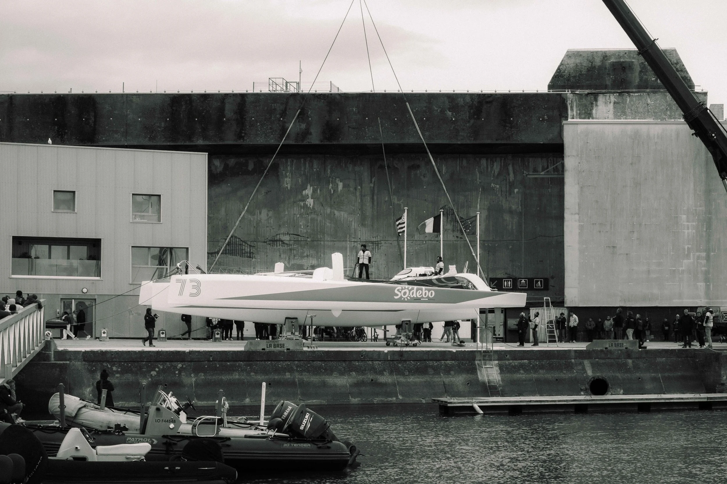 Un bateau de course suspendu en exposition devant un mur en béton, avec des gens autour et un petit bateau pneumatique dans l'eau en premier plan.