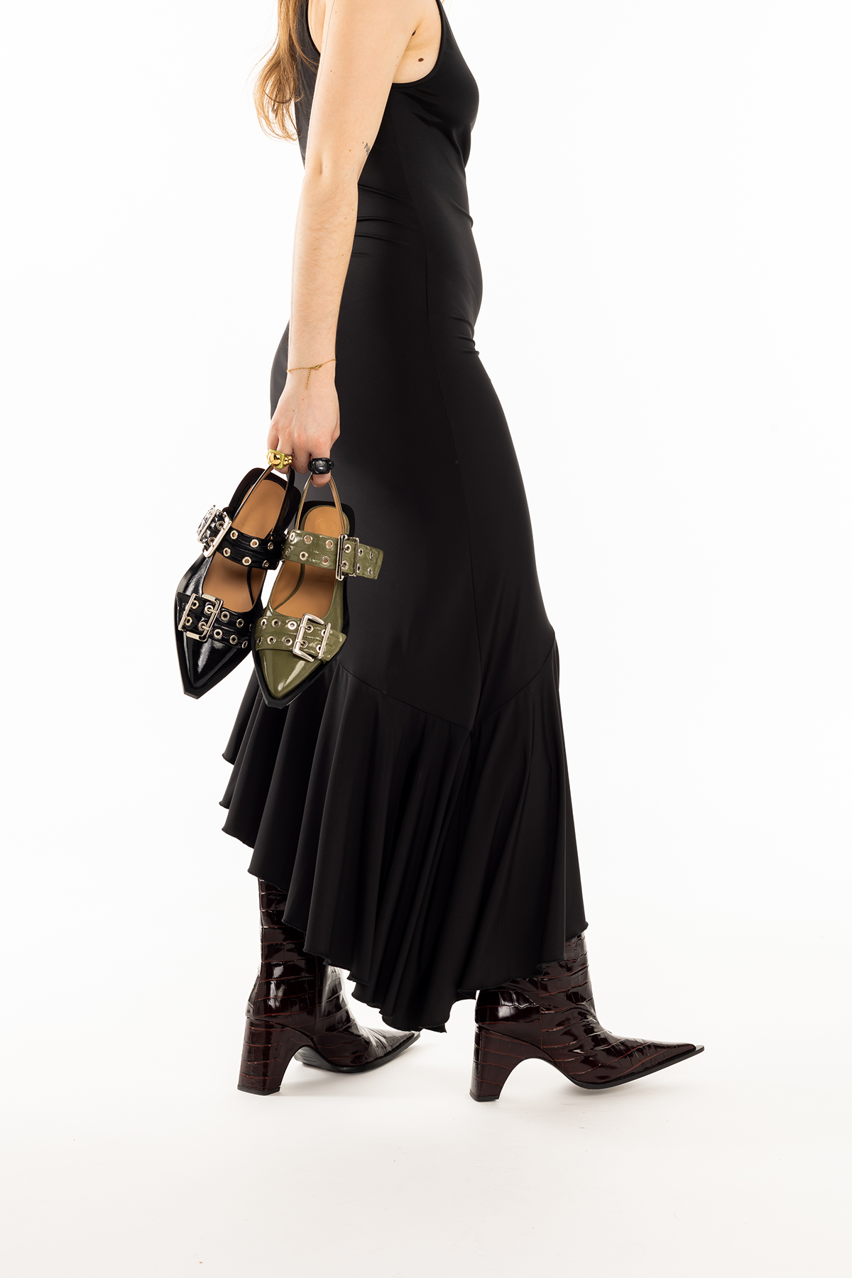 Femme tenant deux paires de chaussures à talons, une noire avec des détails en métal argenté et une vert olive avec des détails en métal argenté, portant une robe noire longue avec des bottines en cuir marron avec un talon épais.
