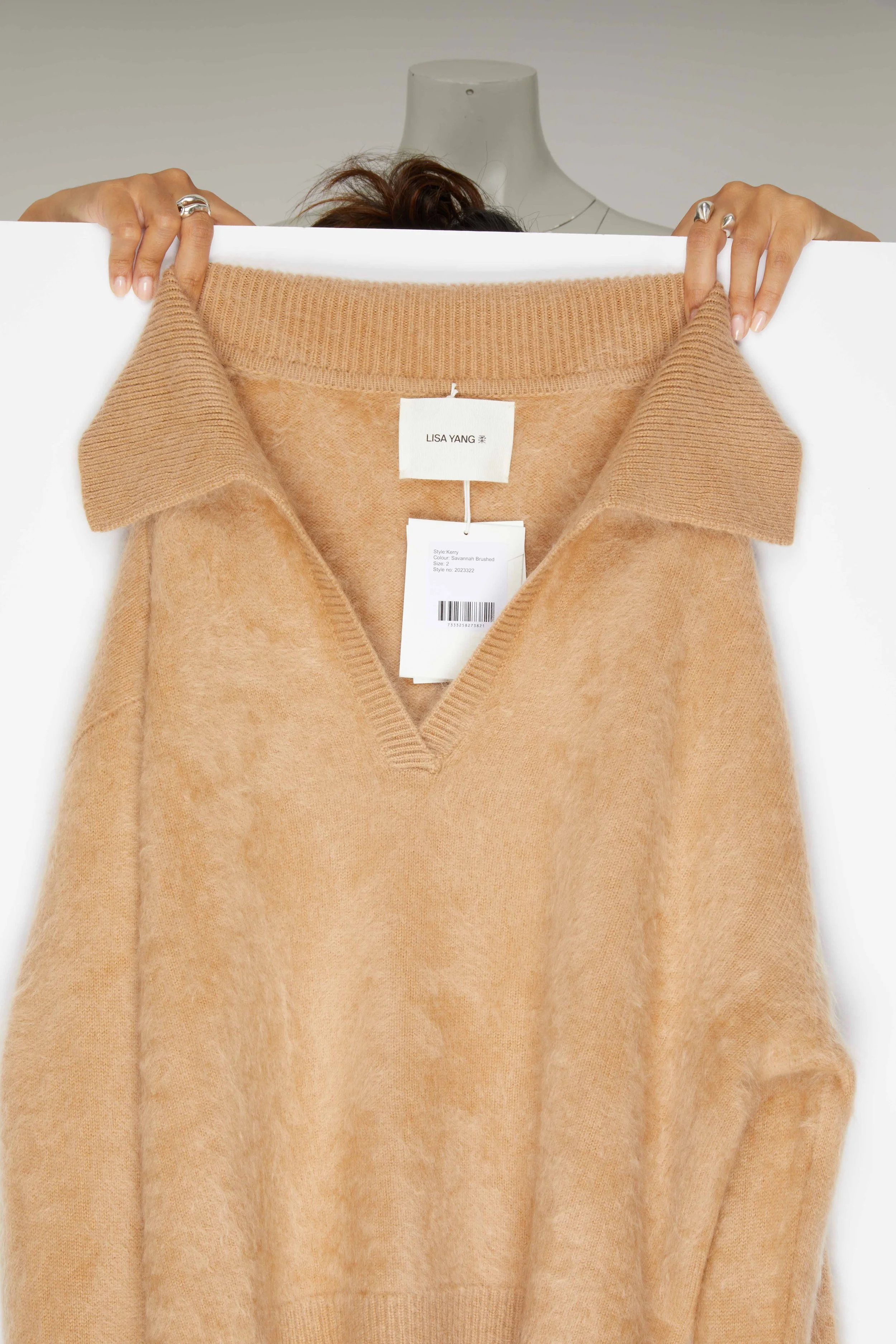 Pull en laine beige sur un mannequin avec des mains tenant une planche blanche en arrière-plan.