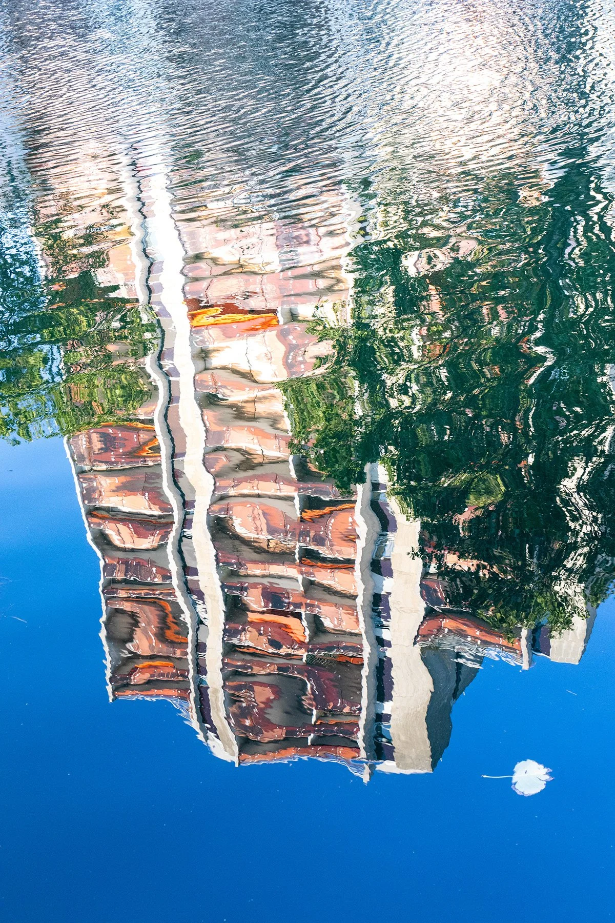 Reflet de bâtiments résidentiels colorés et d'arbres dans l'eau d'une rivière ou d'un canal, avec une barque blanche flottant à la surface.