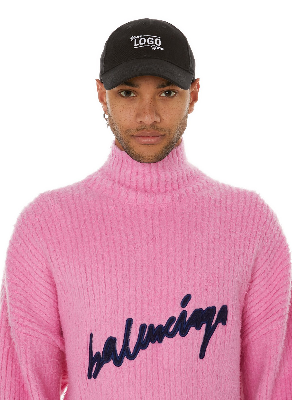 Un jeune homme porte un bonnet noir avec un logo blanc, un pull rose en maille avec le mot 'balenciaga' brodé en lettres bleues, et regarde vers l'objectif.