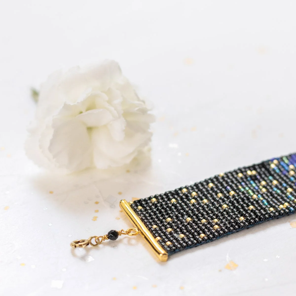 Un bracelet en perles noires et dorées avec un fermoir en or, posé sur une surface blanche avec une fleur blanche en arrière-plan.
