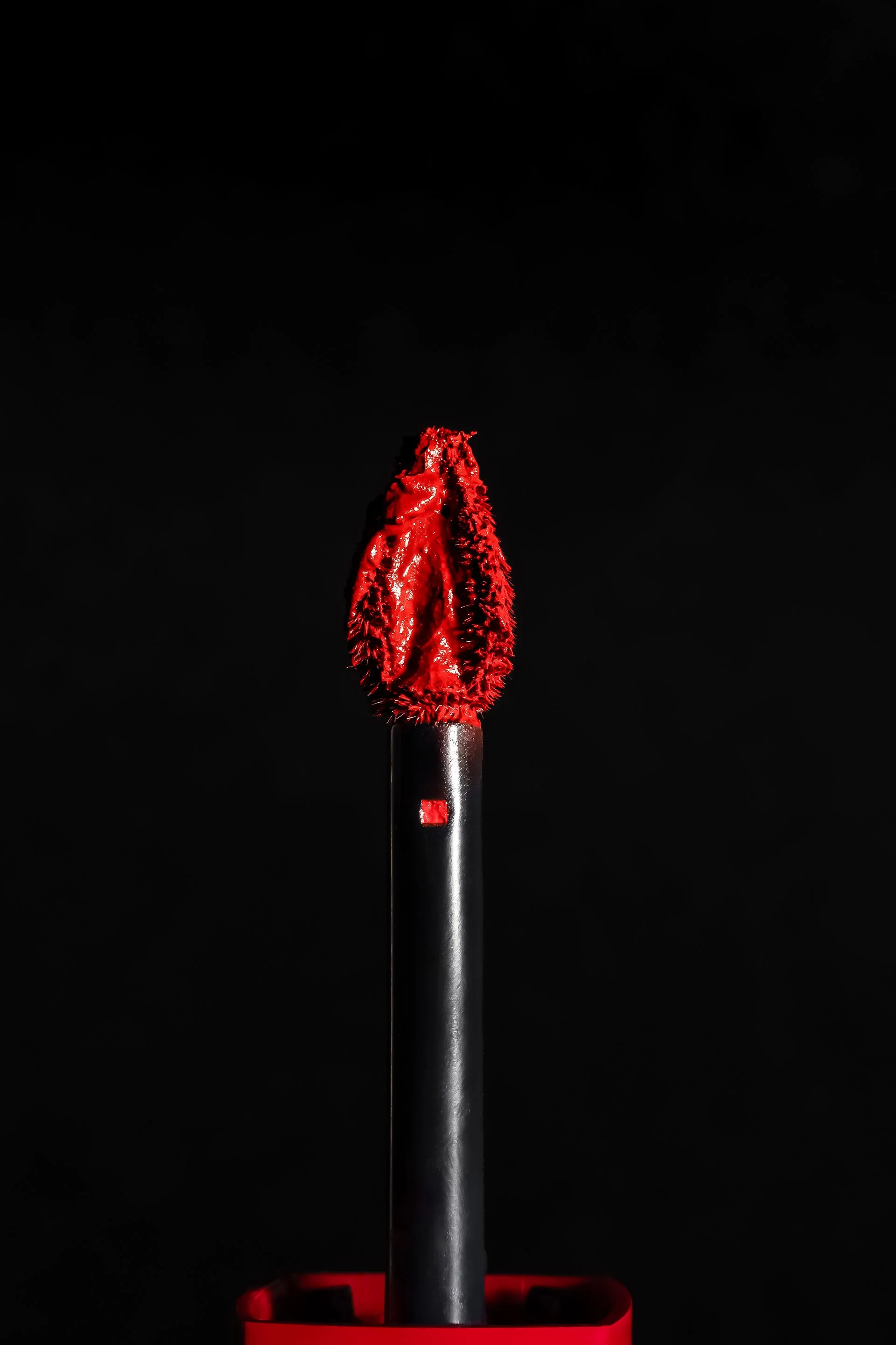 Un pinceau de maquillage avec un embout en forme de crâne rouge, placé dans un support rouge, sur fond noir.