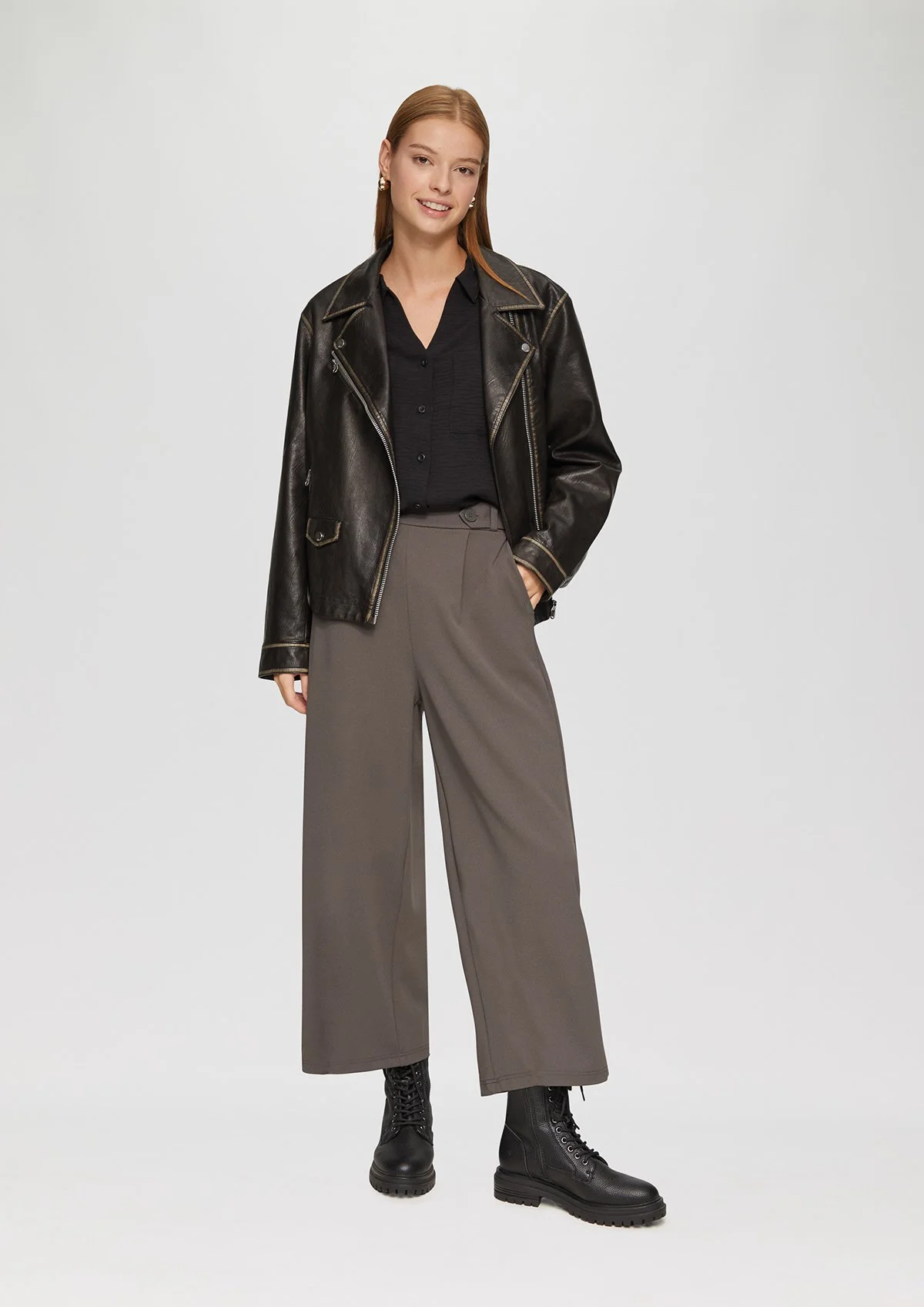 Une jeune femme portant une veste en cuir noir, un pantalon large gris et des bottes noires, se tenant devant un fond gris clair.