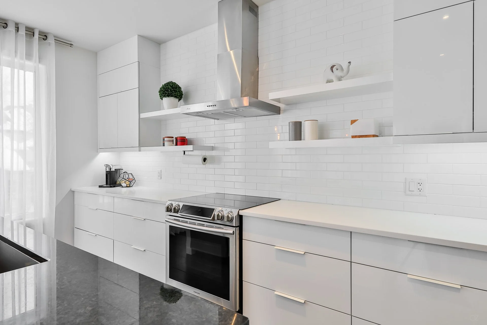 Cuisine moderne avec des armoires blanches, un plan de travail blanc, un four électrique en acier inoxydable, une hotte aspirante en acier inoxydable, des étagères blanches avec des décorations, et des fenêtres avec rideaux blancs.