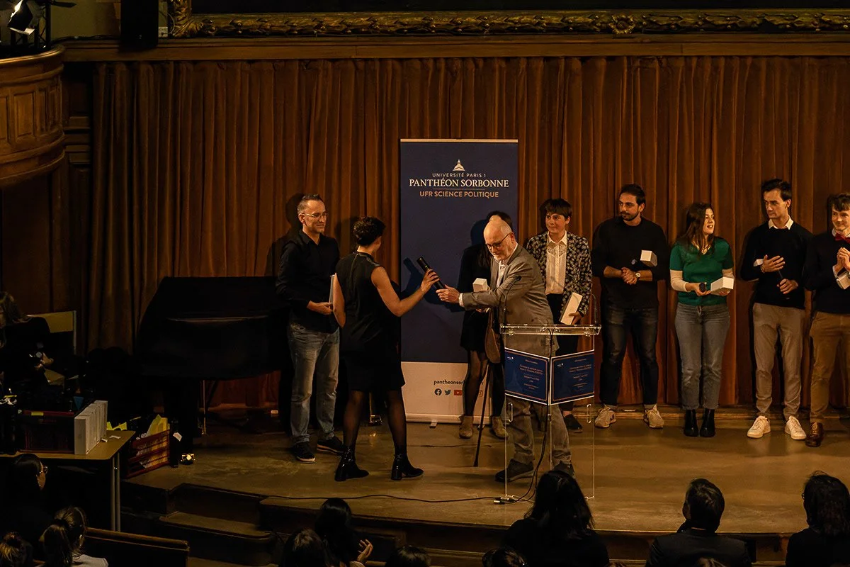 Cérémonie ou remise de prix à l'université Paris 1 Panthéon Sorbonne, avec plusieurs personnes sur scène, dont une femme recevant un trophée ou un prix, et un homme en train de lui remettre le trophée, dans un auditorium avec un fond en bois et un pa