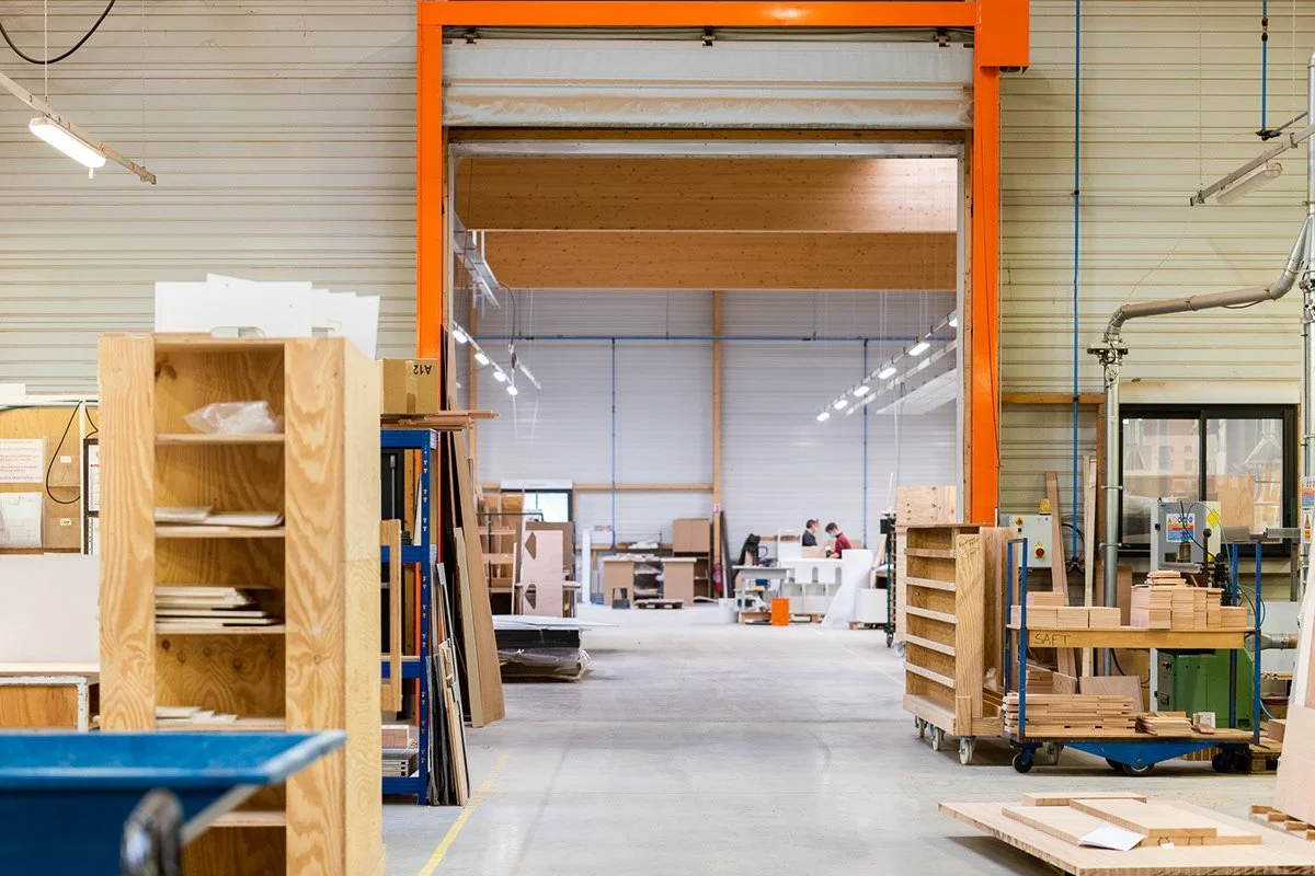 Intérieur d'une usine de fabrication de meubles en bois avec étagères, chariots en bois, travailleurs au fond et équipement de travail.