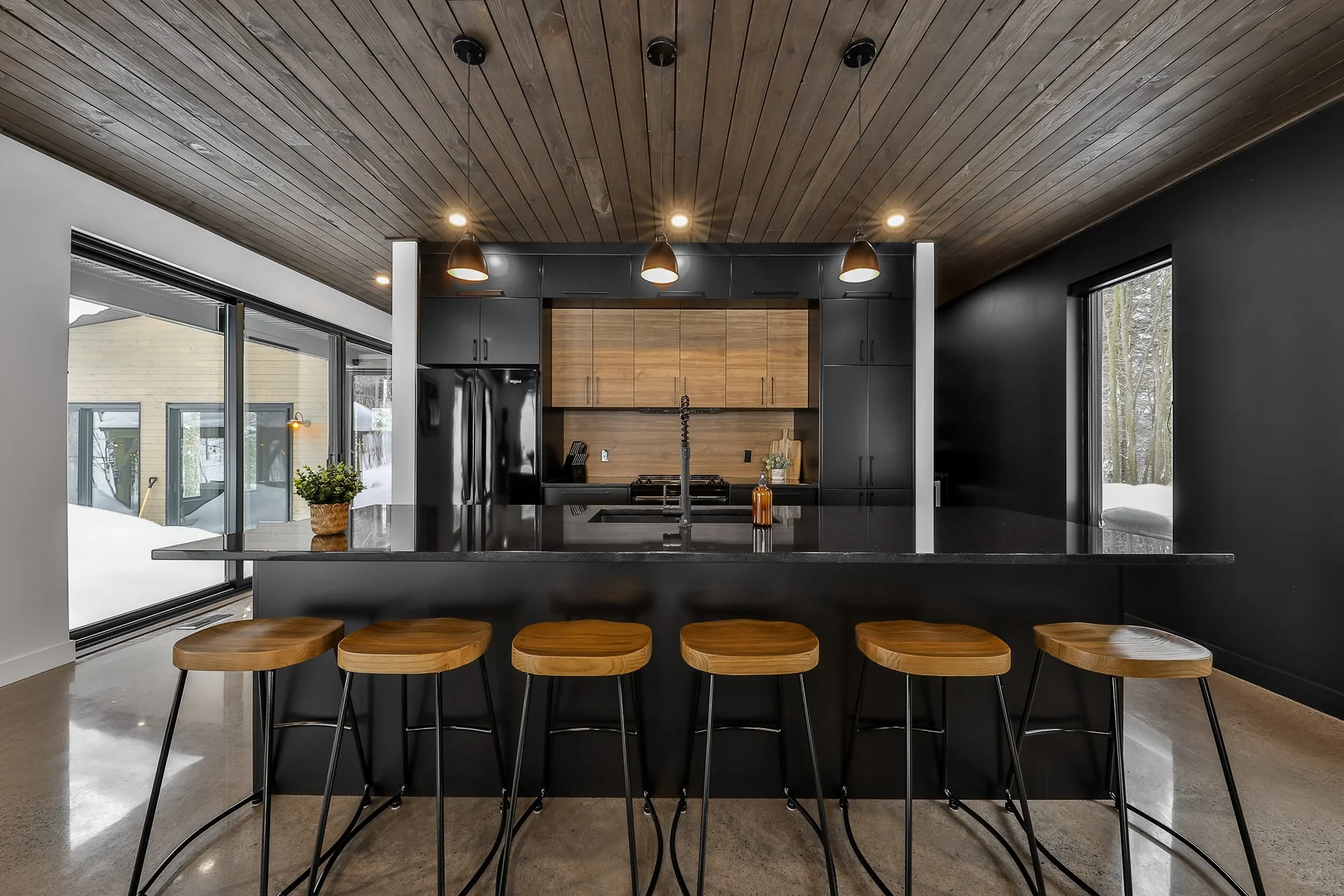 Cuisine moderne avec plan de travail noir, armoires noires et bois, fenêtres de grande taille, et cinq tabourets en bois.
