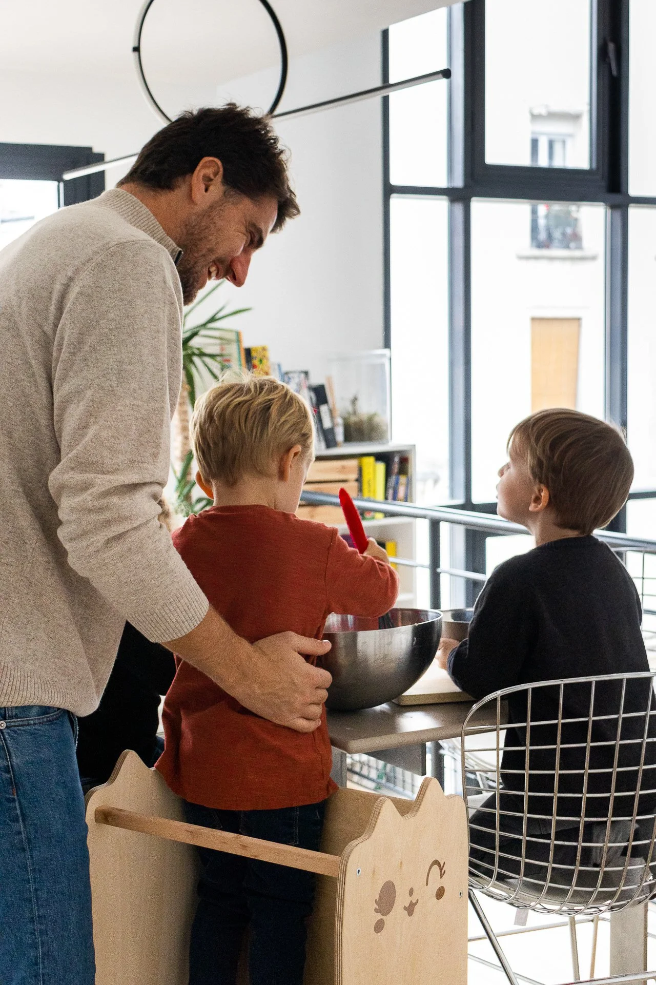 Un homme souriant regarde deux enfants qui cuisinent ou jouent ensemble à une table dans une cuisine moderne et lumineuse.