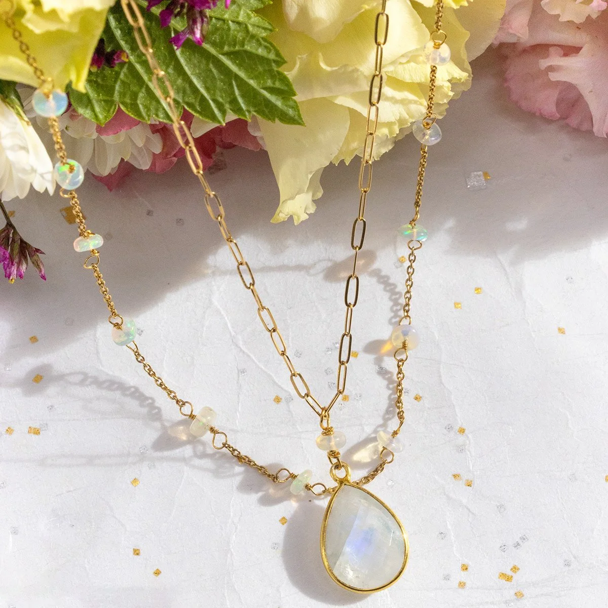 Collier avec pendentif en pierre lunaire entouré d'une chaîne en or, décoré de petits cristaux et posé sur une surface blanche avec des fleurs en arrière-plan.