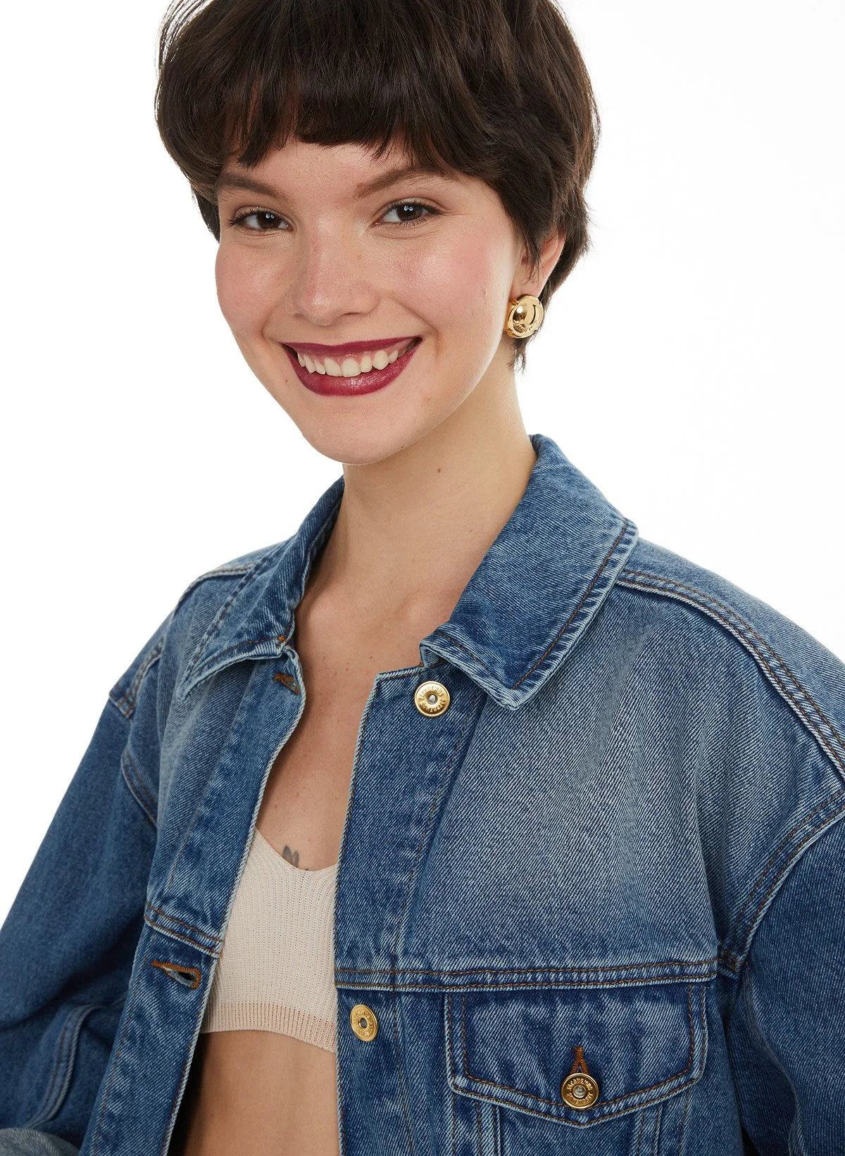 Jeune femme avec coupe courte brune, souriante, portant un jean bleu et une veste en denim avec boutons dorés, fond blanc.
