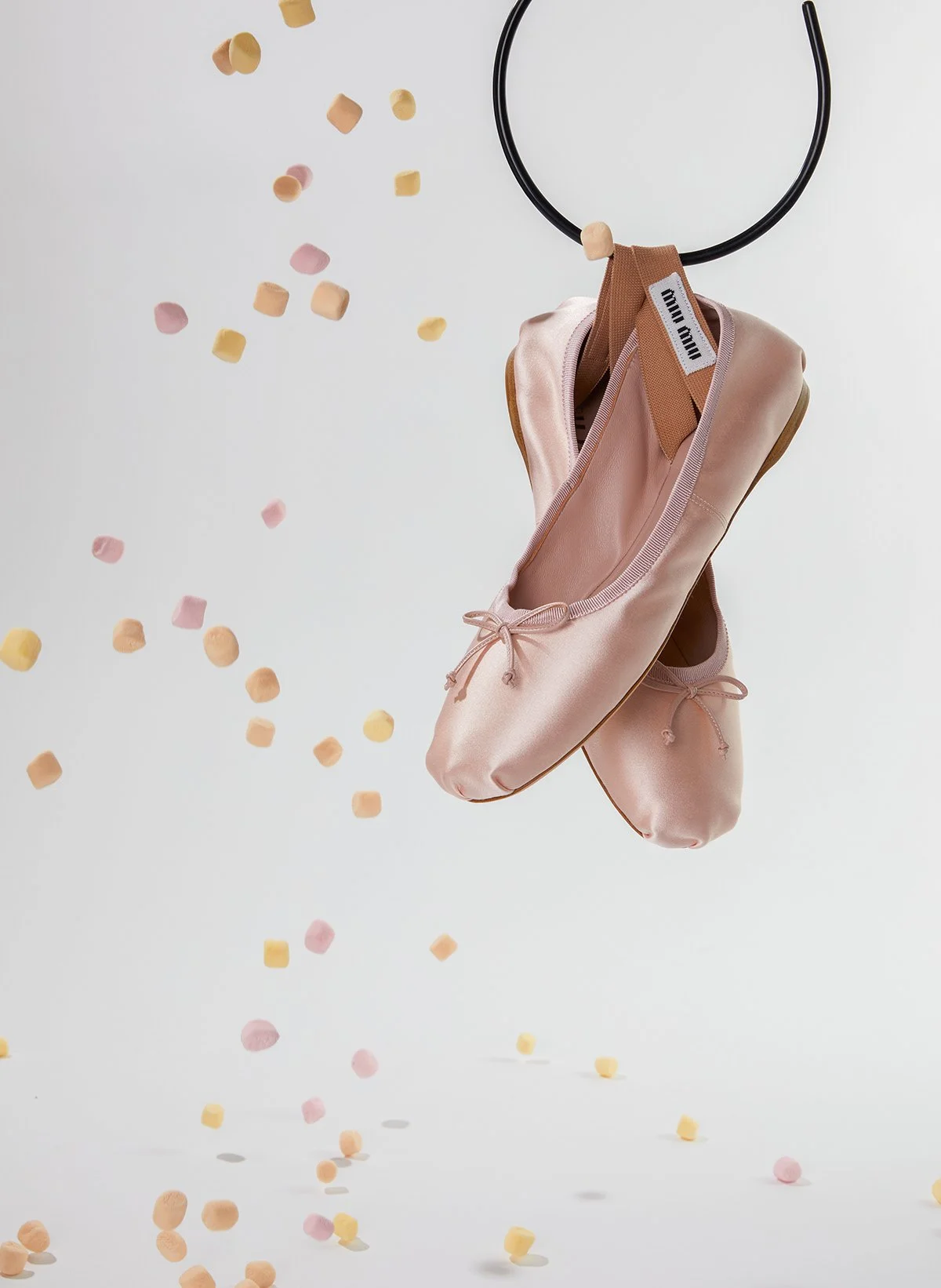 Une paire de chaussons de ballet rose pâle suspendus à un anneau noir, avec des confettis de couleur pastel tombant autour sur un fond blanc.