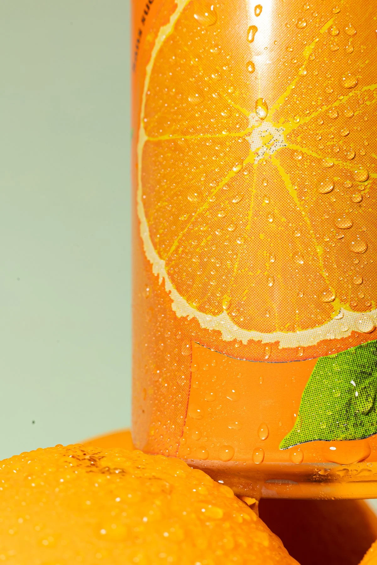 Une canette de boisson fruitée à motif d'orange, avec une illustration d'une tranche d'orange et une feuille verte, humide avec des gouttes d'eau, et un citron en dessous.