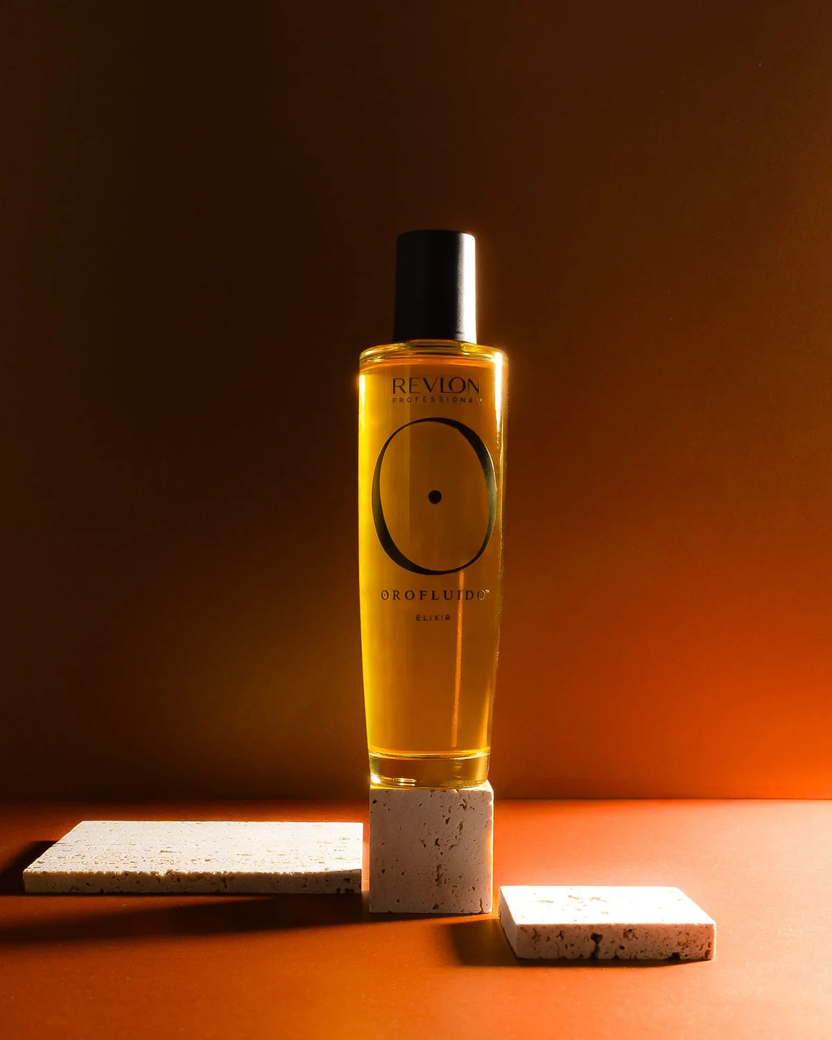 Une bouteille de parfum Orofluido Elixir posée sur un bloc blanc, avec un fond chaud et un éclairage ciblé.