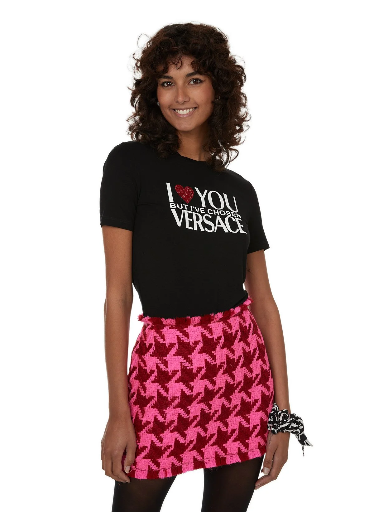 Une femme souriante porte un t-shirt noir avec la phrase 'I ❤️ YOU BUT I'VE CHOSEN VERSACE' et une jupe rose à motifs houndstooth noir.
