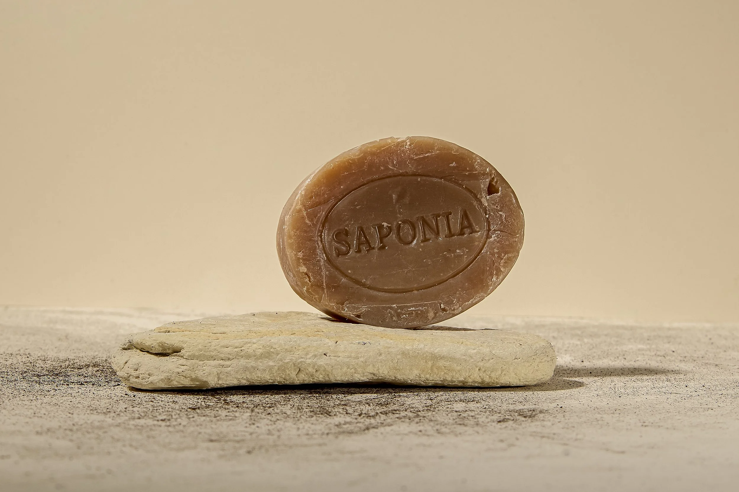 Un savon marron avec l'inscription 'SAPONA' posé sur une pierre plate, sur une surface de sable ou de poussière, avec un fond neutre beige.
