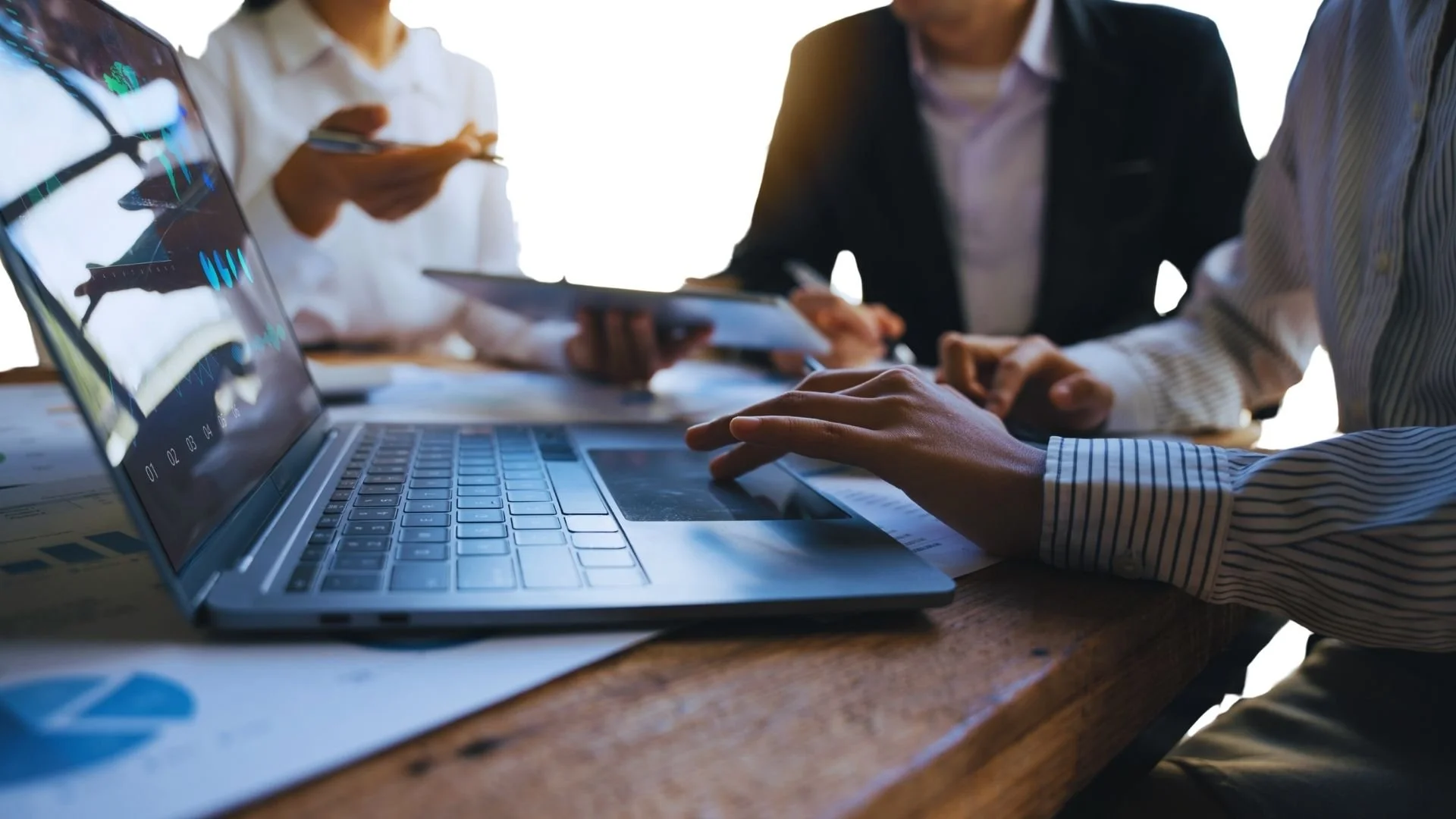 People in business attire working around a laptop on a wooden table, discussing documents and digital data.