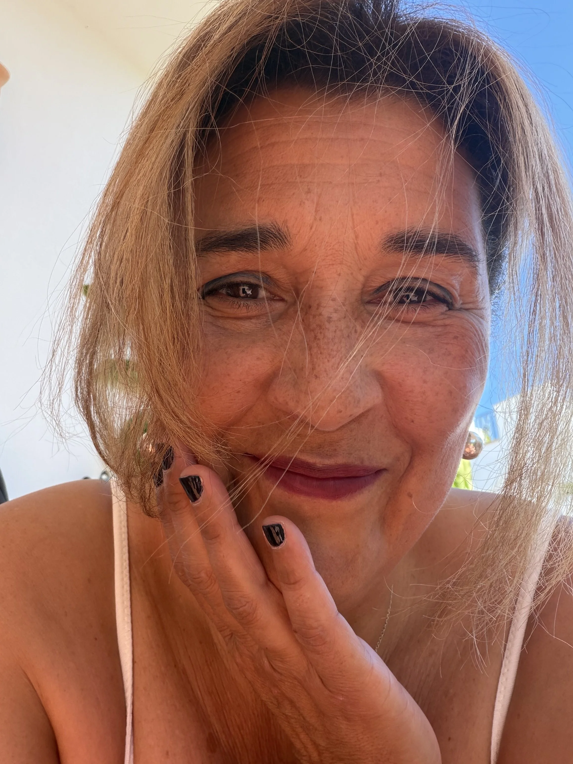 Close-up of Sandra Teale with light brown hair and a big smile, touching her face with her right hand, wearing a white tank top.