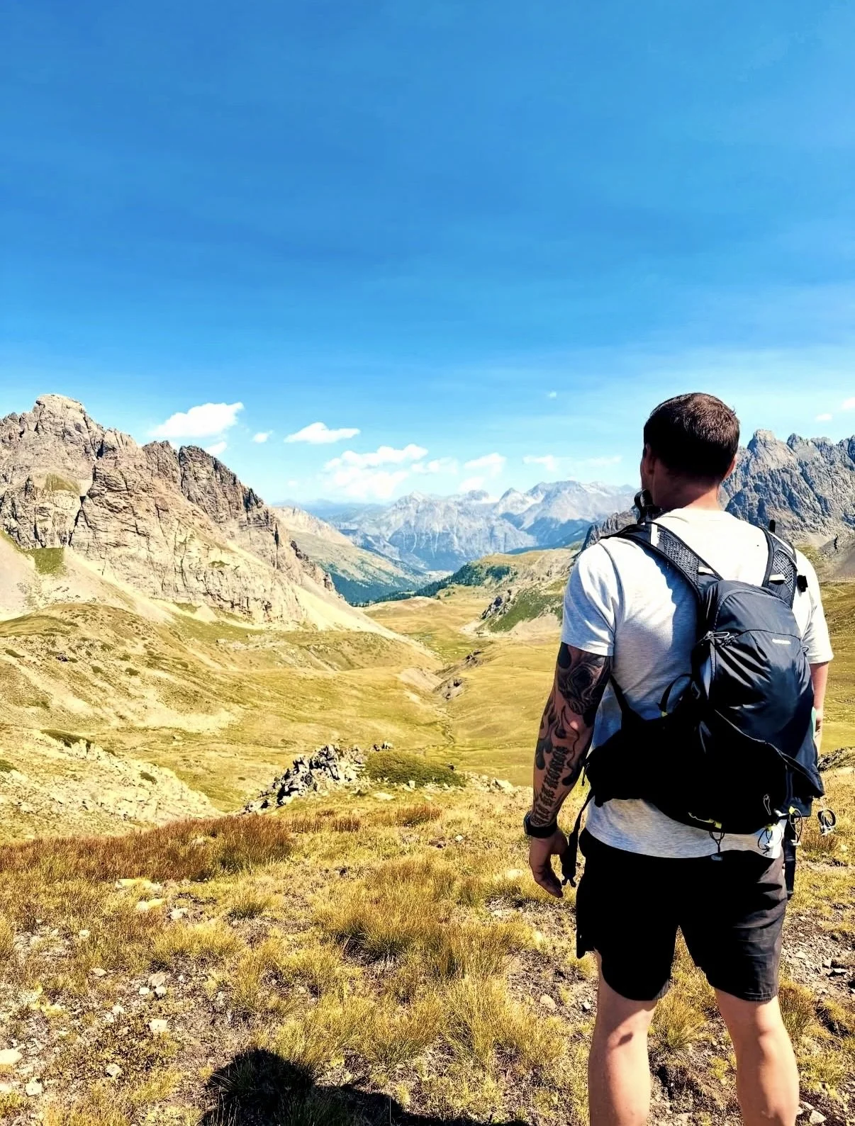 Un homme avec un sac à dos, regardant un paysage de montagnes et de vallées ensoleillées sous un ciel bleu.