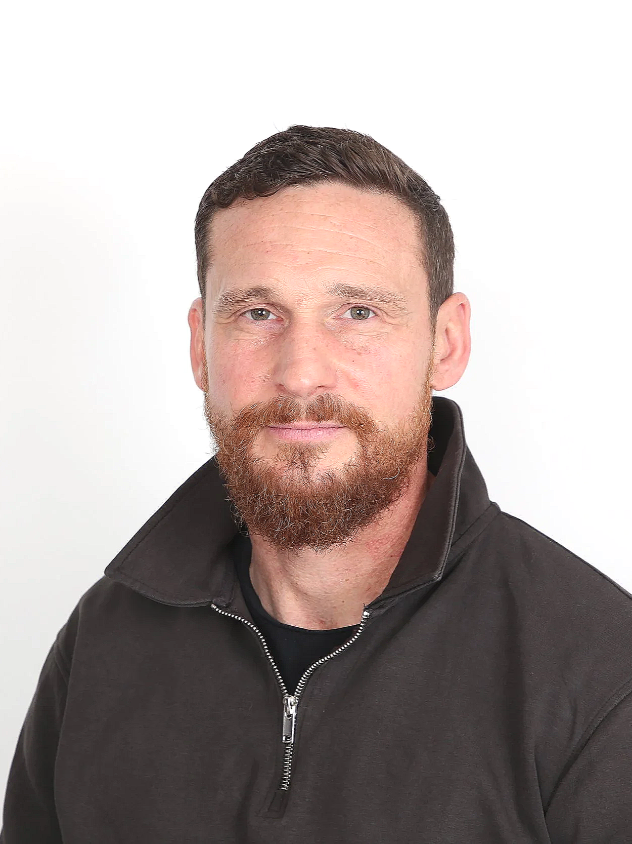 Portrait d'un homme avec une barbe brune, portant un sweat noir avec fermeture éclair, fond blanc.