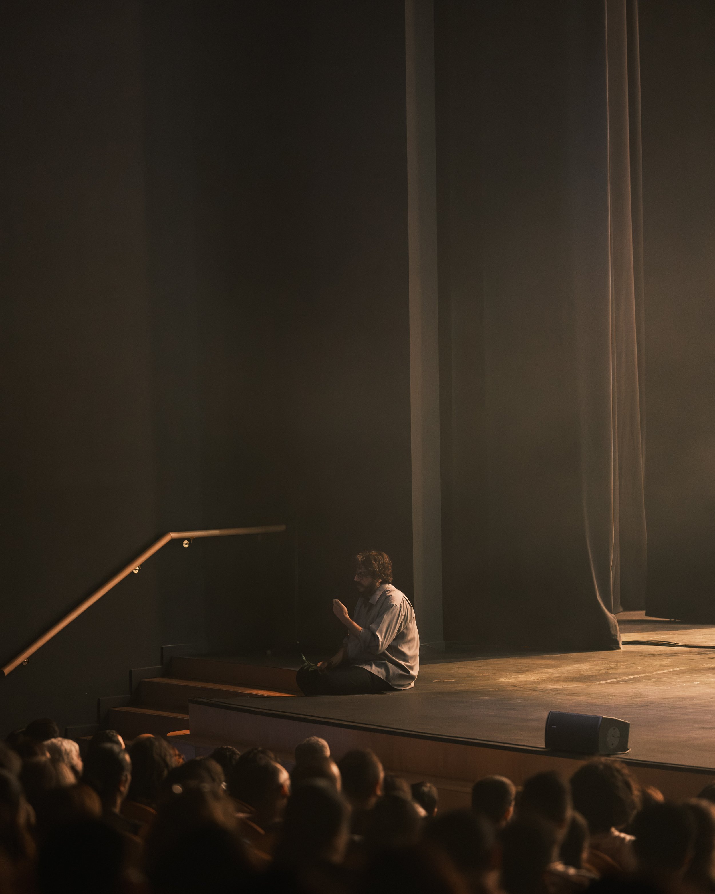 A man sitting on a stage, using his phone, with an audience in front of him and a large dark curtain backdrop.