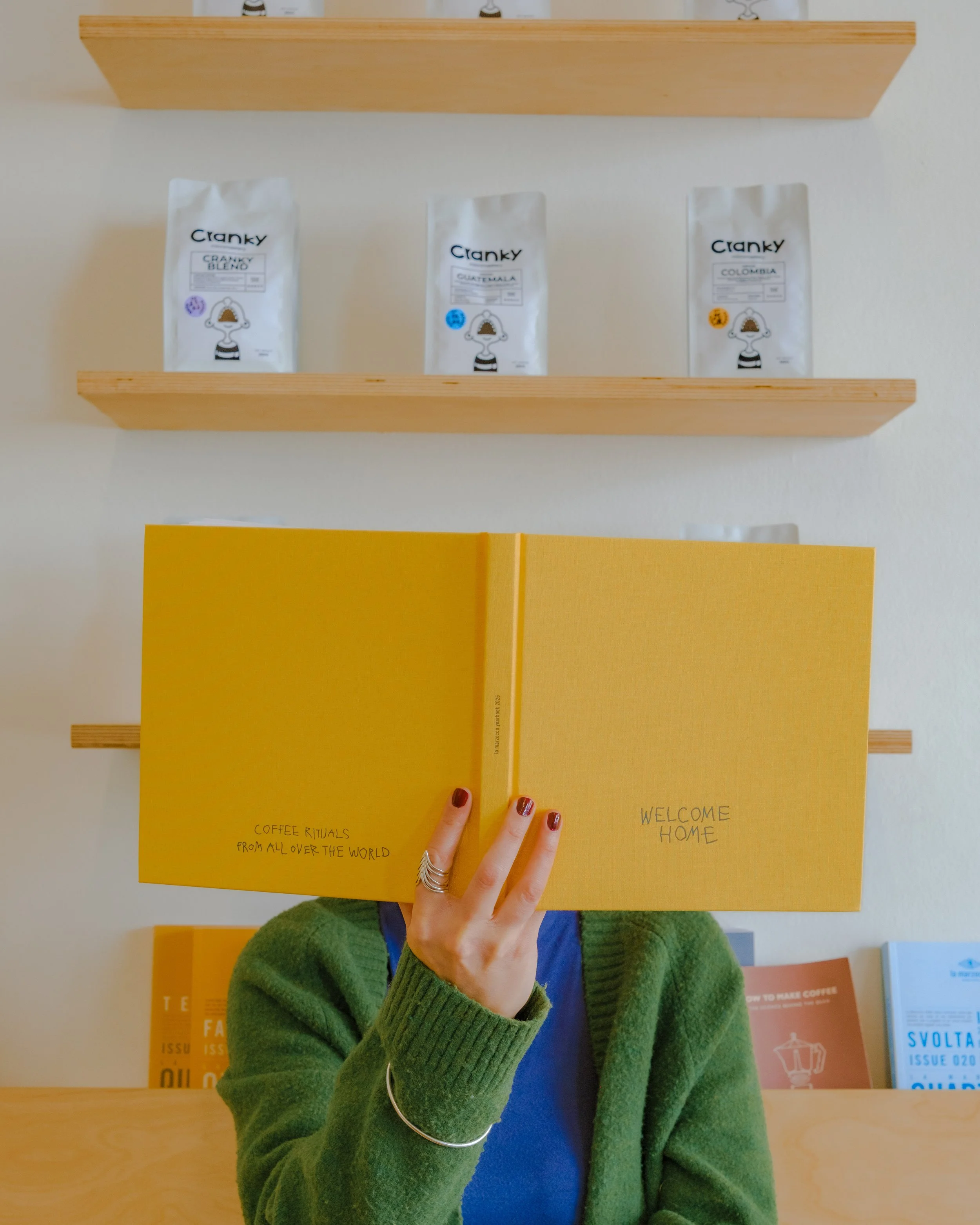 Person holding yellow book in front of face, wearing green sweater, blue shirt, with shelves of colorful magazines or posters behind.